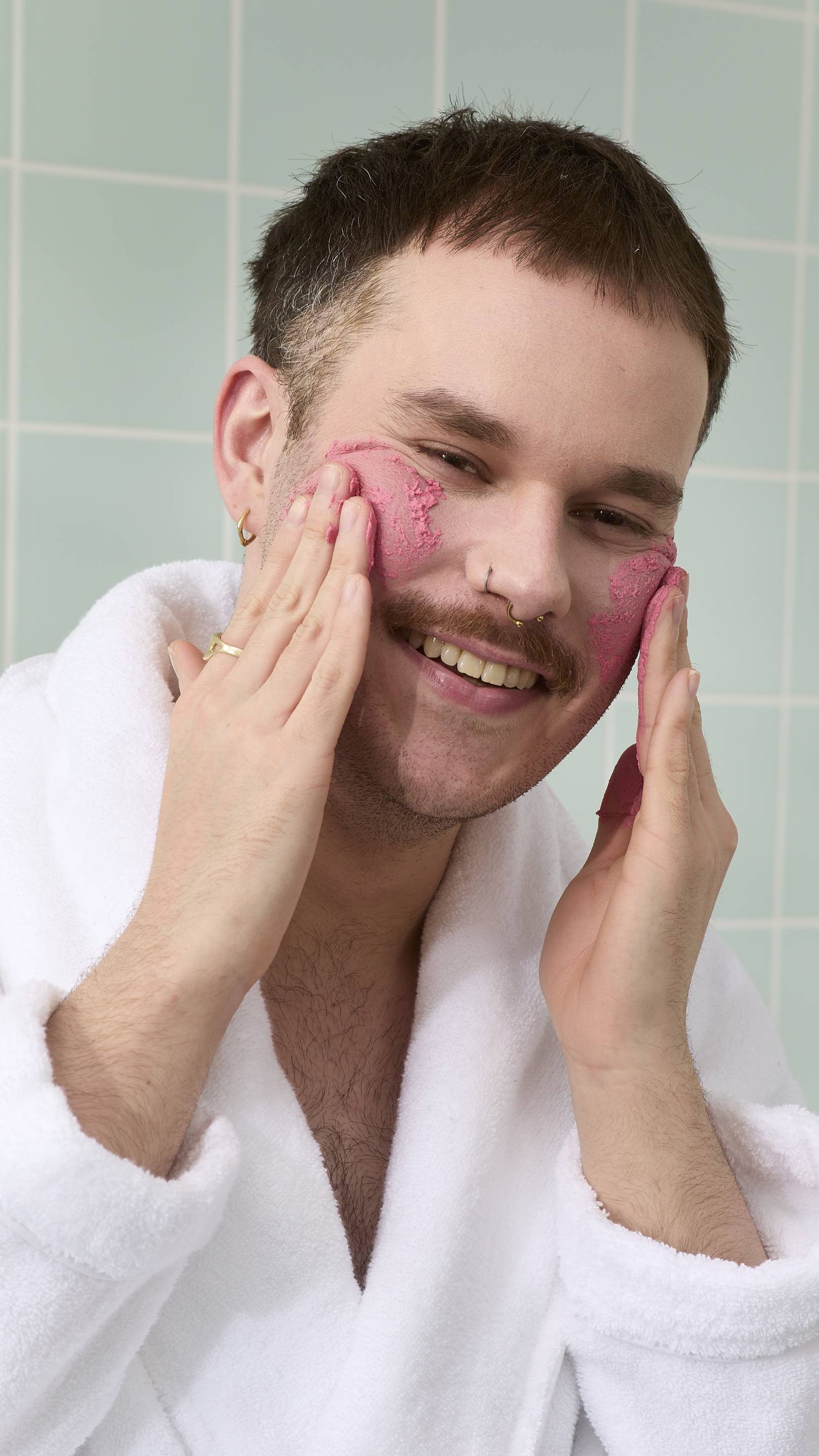 A close-up of a smiling model with a moustache and facial piercings wearing a white dressing gown. They are applying a textured pink "Ladybird" face cleanser to their cheeks with their fingertips against a background of light green bathroom tiles.