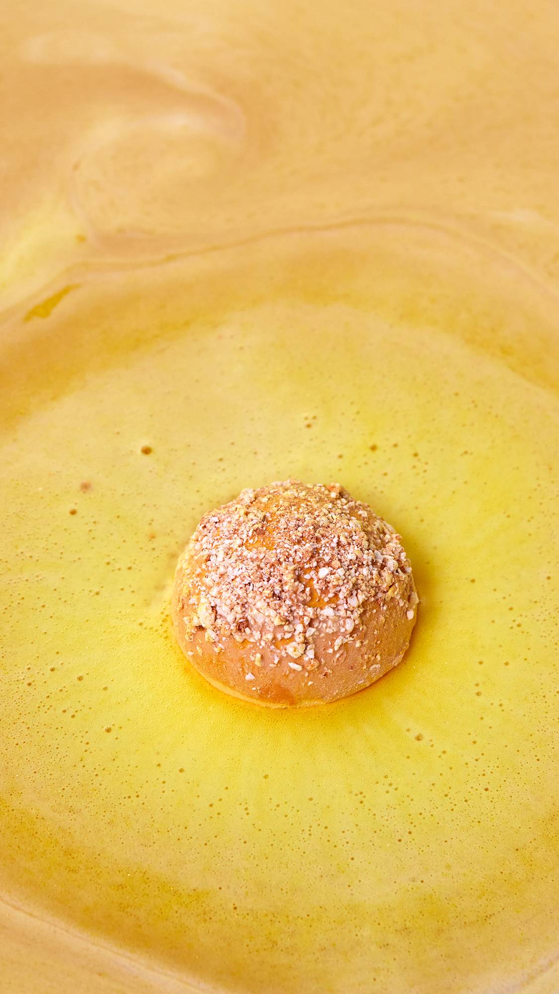 An aerial image of the Let The Good Times Roll bath bomb as it starts to dissolve in the bath. The product gives off thick, bubbling, caramel-coloured foam across the surface with flecks of golden glitter throughout. 