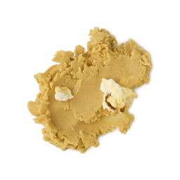 A close-up, overhead view of a thick, crumbly smudge of product (likely a scrub or balm) in a warm tan or yellow-gold color. Two small, lighter creamy white pieces are embedded in the texture, all set against a light grey background.