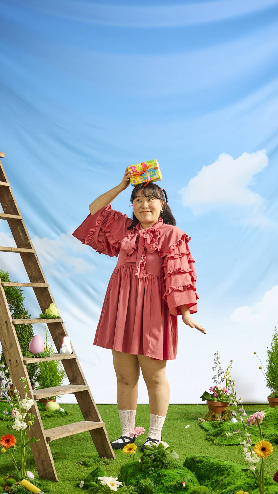 The model is standing in a ruffled coral dress under blue skies, surrounded by grass and flowers, holding the gift box on top of their head. 