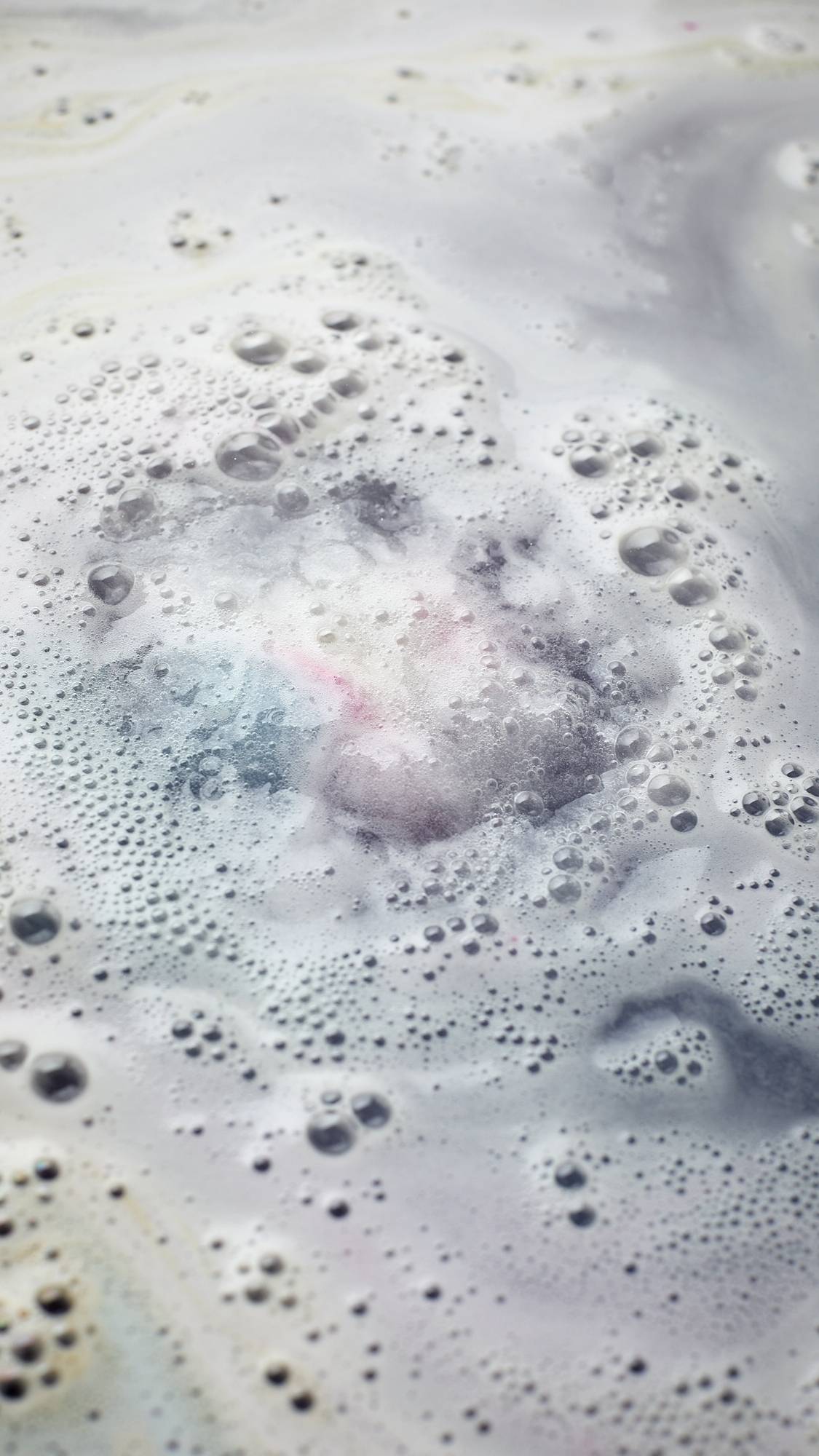 The image shows a view of the bath water from above after using the Lush, Liberation, blue, pink and white bath bomb. The product has fully dissolved leaving behind a delicate layer of velvety foam on the surface of the water. 