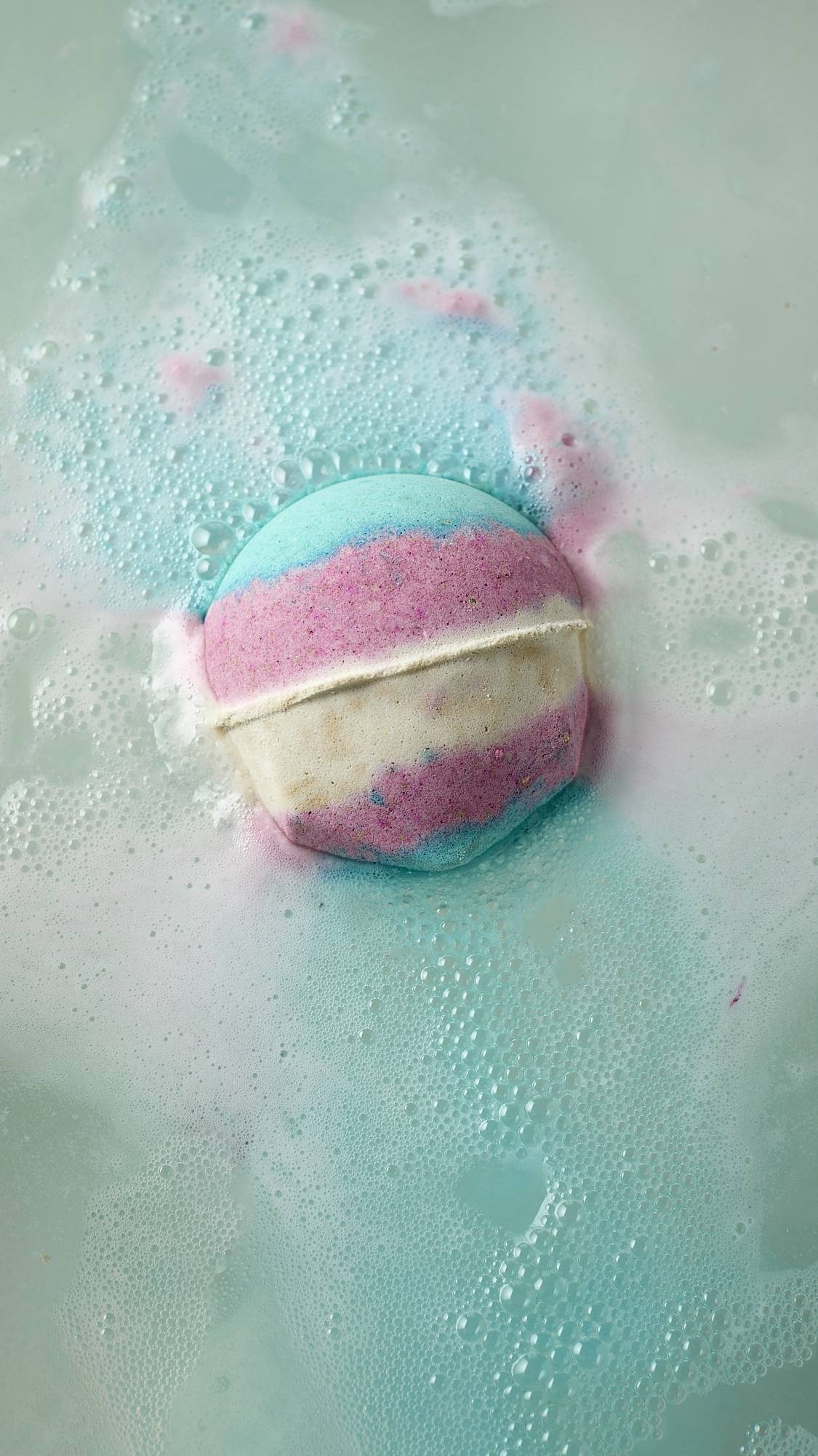 The image shows an aerial view of the blue, pink and white, Lush, Liberation bath bomb as it begins to dissolve in the bath water. The product is giving off a delicate layer of sky-blue foam. 