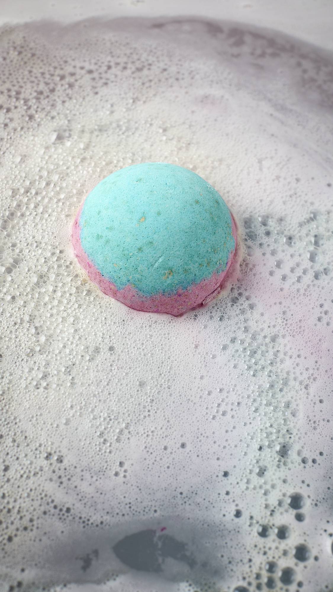 The image shows an aerial view of the blue, pink and white, Lush, Liberation bath bomb as it begins to dissolve in the bath water. The product is giving off a delicate layer of sky-blue foam. 