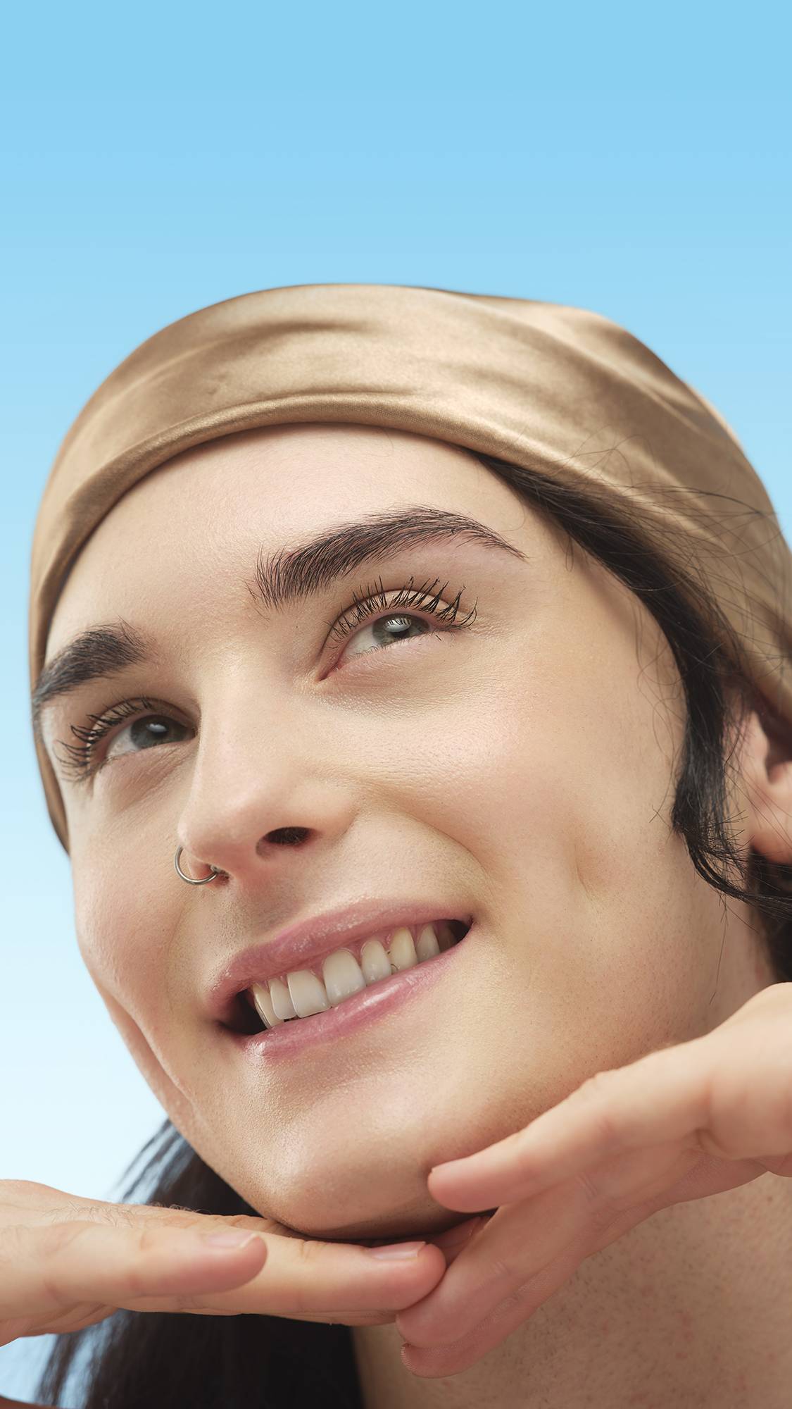 A close-up portrait of a person with neutral skin, wearing a scarf and nose ring. They smile, cupping their chin, with the Light Medium Neutral Beauty Balm on their face, against a blue background.