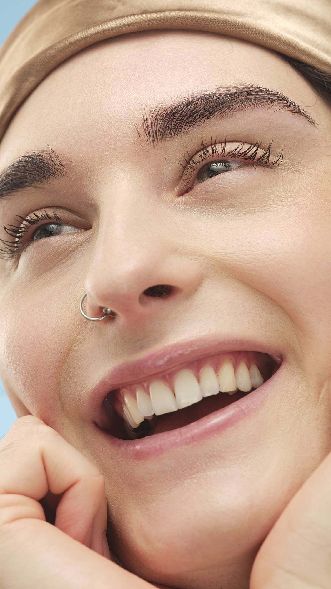 A close-up vertical shot of a smiling person's face, looking up, with a hand resting on their chin. They wear a nose ring and a satin head scarf. The Light Medium Neutral Beauty Balm provides smooth coverage to their light-medium skin.