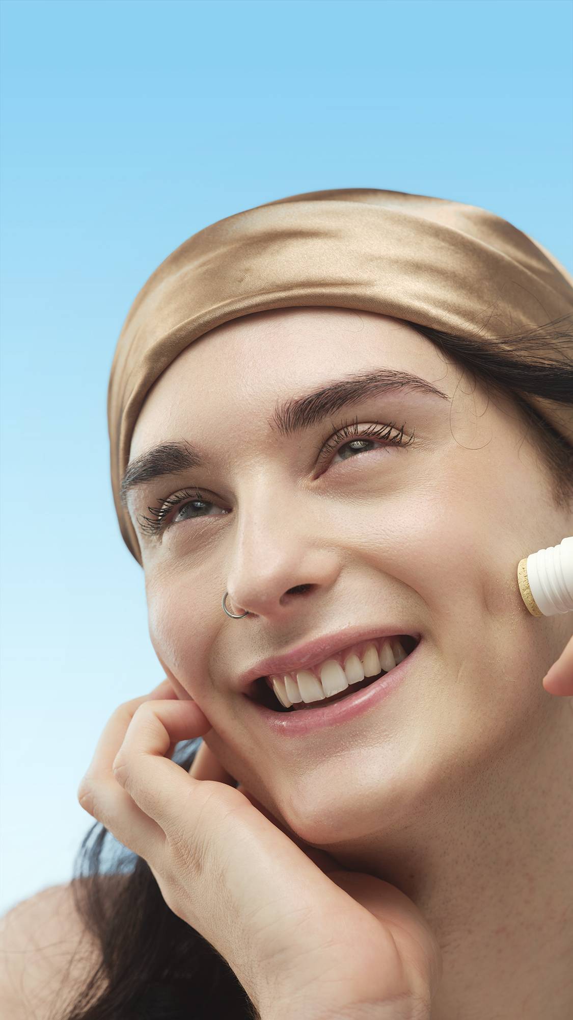 A close-up portrait of a smiling person with neutral skin, wearing a scarf and nose ring. They look up, applying the Light Medium Neutral Beauty Balm stick to their cheek, against a blue background.