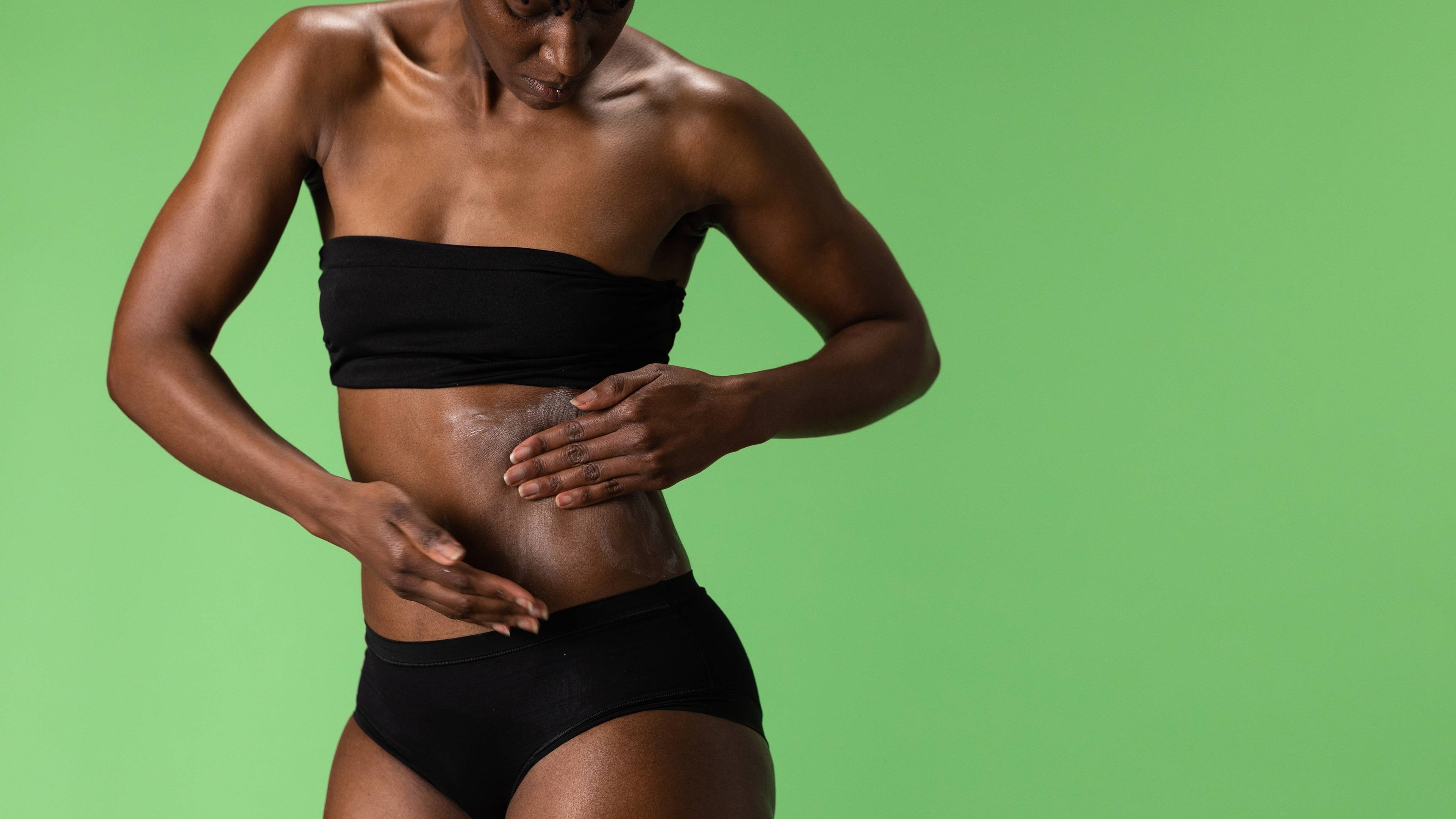 The model is on a fern-green background wearing a black bandeau bra and underwear as they apply the Lime Bounty body butter to their side and stomach area. 