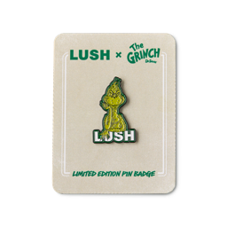 A Lush x The Grinch limited edition enamel pin badge featuring The Grinch resting his chin on his hand above the Lush logo, mounted on a cream backing card