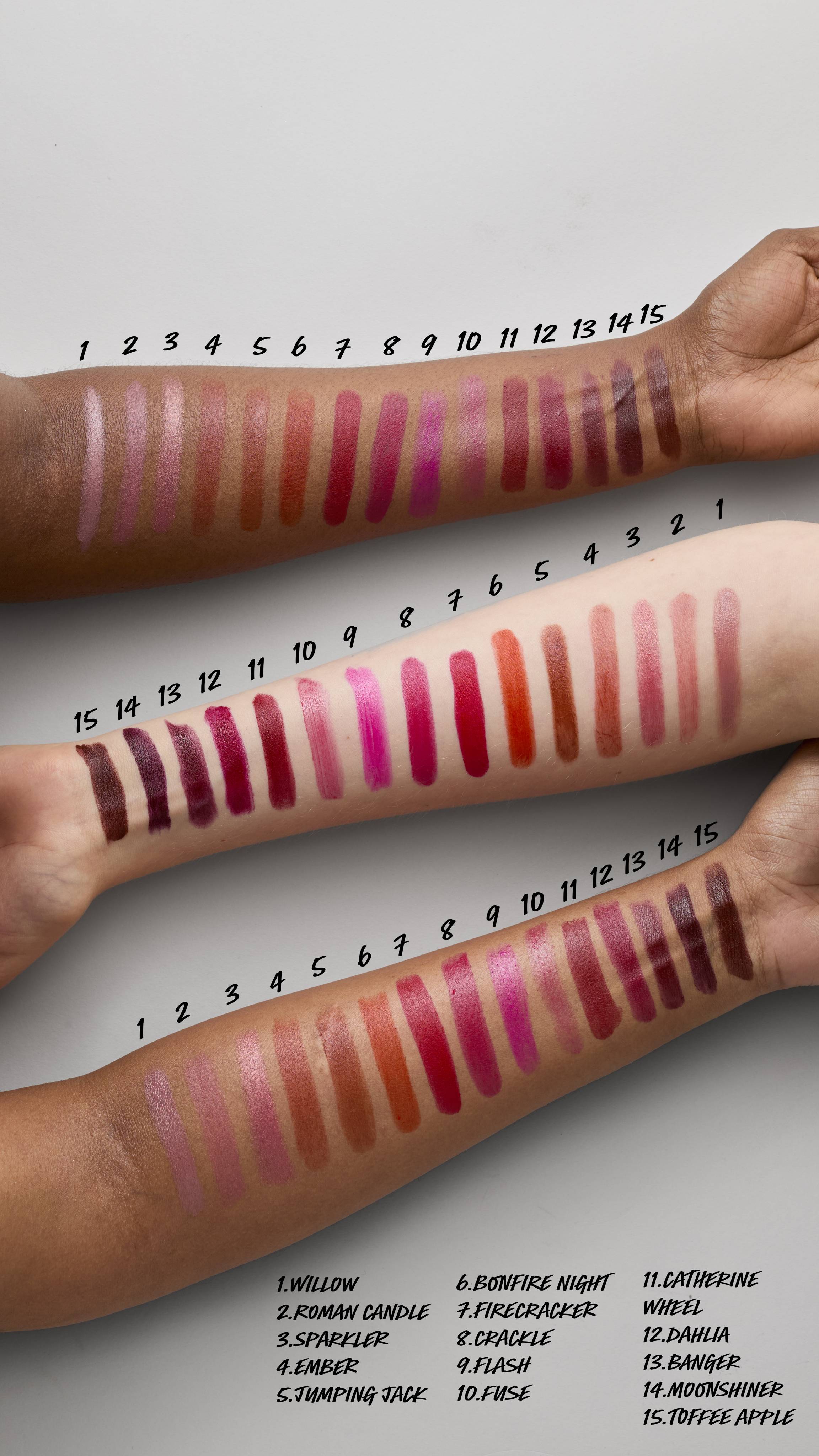 The image shows an aerial view of three forearms of differing skin tones. Across each of the arms are swatched the fifteen Firework Lush lipstick shades, from light to dark, to display how they show on different skin tones. 