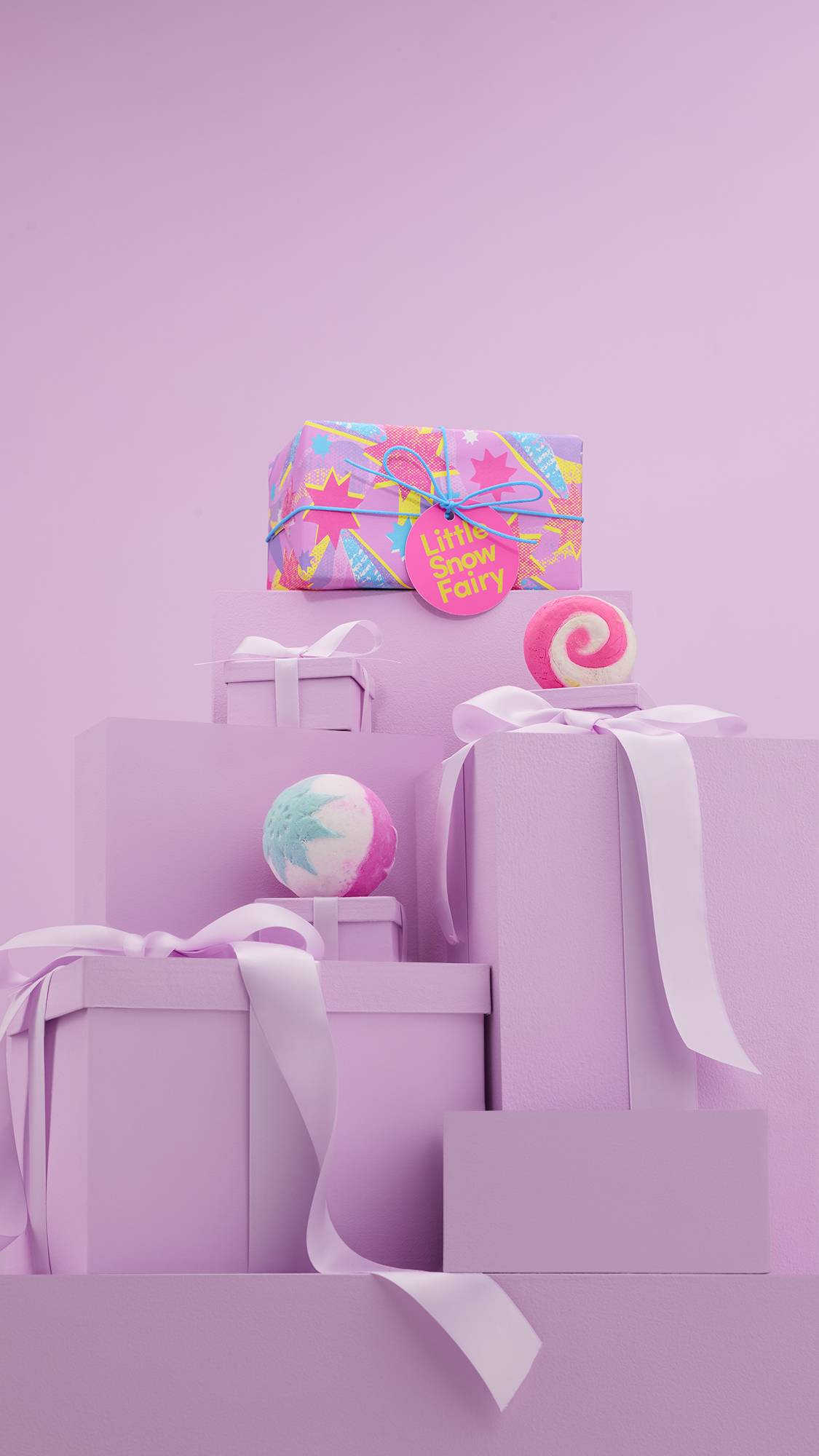 The image shows a large pile of differently-sized wrapped pink gifts with ribbons, on a matching pink background. The vibrant Little Snow Fairy gift box sits on top of the pile, and the two included Lush products sit on other lower pink gifts.