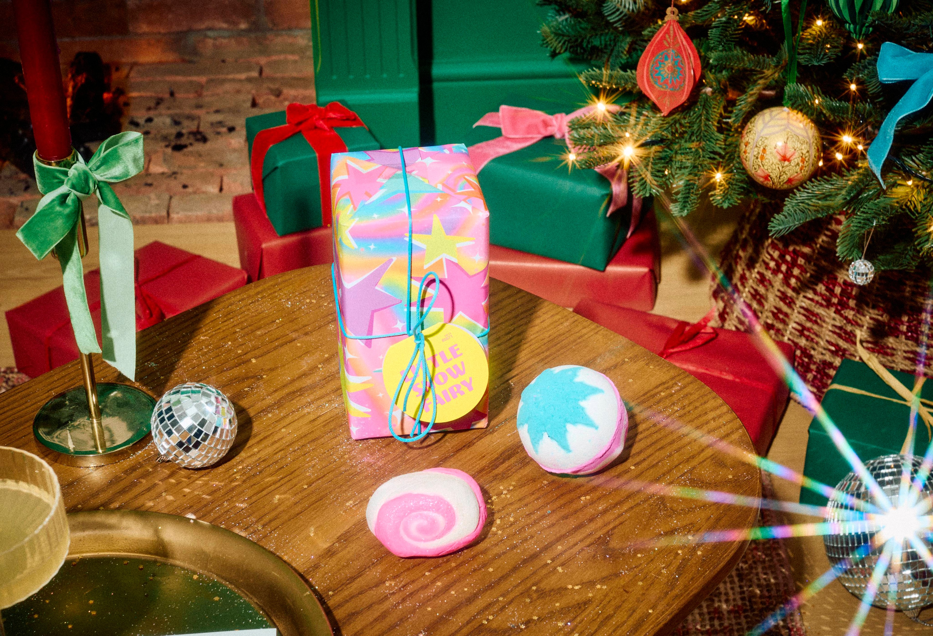The Lush Little Snow Fairy gift set wrapped in colourful starry paper stands on a wooden table alongside a blue snowflake bath bomb and a pink swirled bubble bar, set in a festive room with gifts and a Christmas tree.