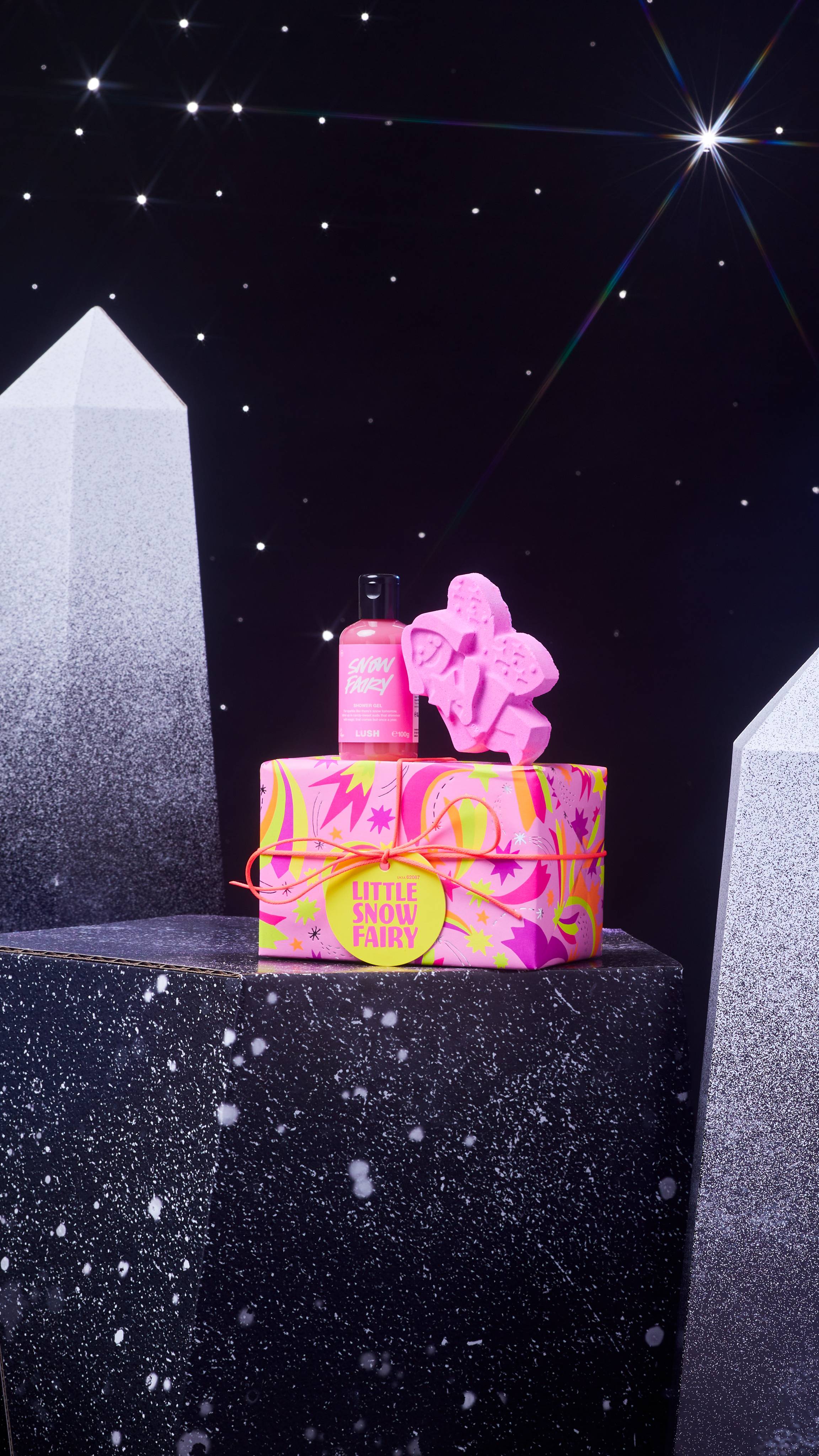The Snow Fairy gift box and two products are on a dark chromatic platform among geometric shapes.