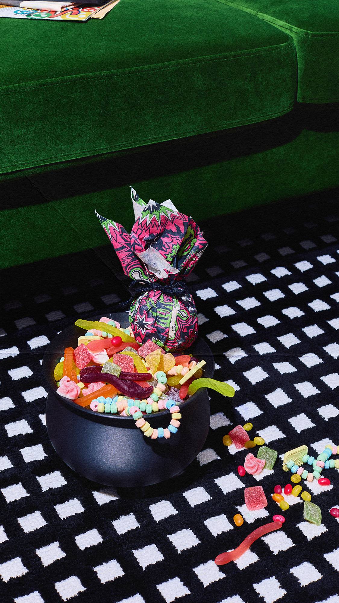 A black cauldron full of colourful jelly sweets and candy necklaces sits next to a small, brightly wrapped gift on a black and white chequered rug. A vibrant green velvet couch is visible in the background.