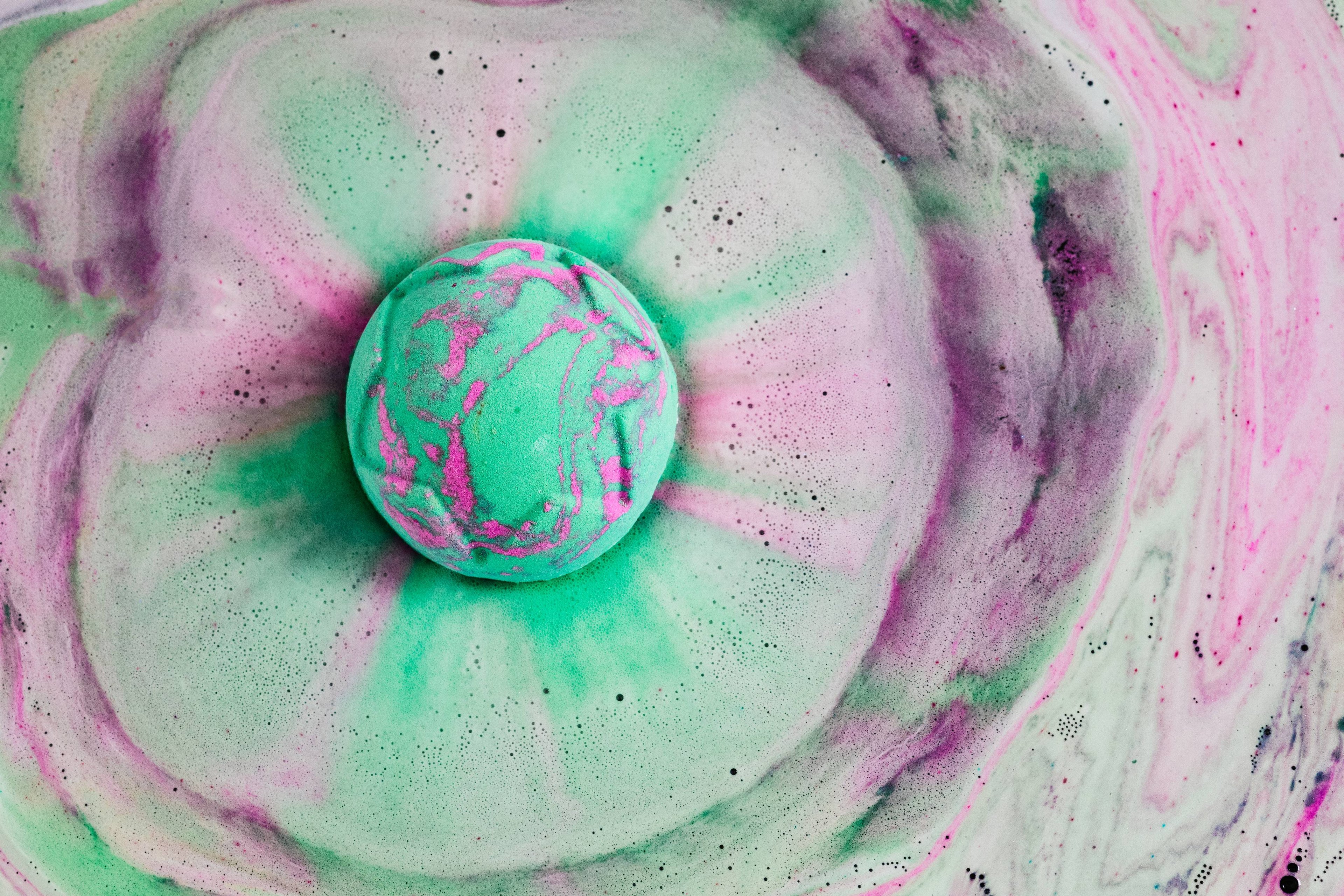 The bath bomb sits on top of the water, dispersing foamy pink and green, frothy swirls. 