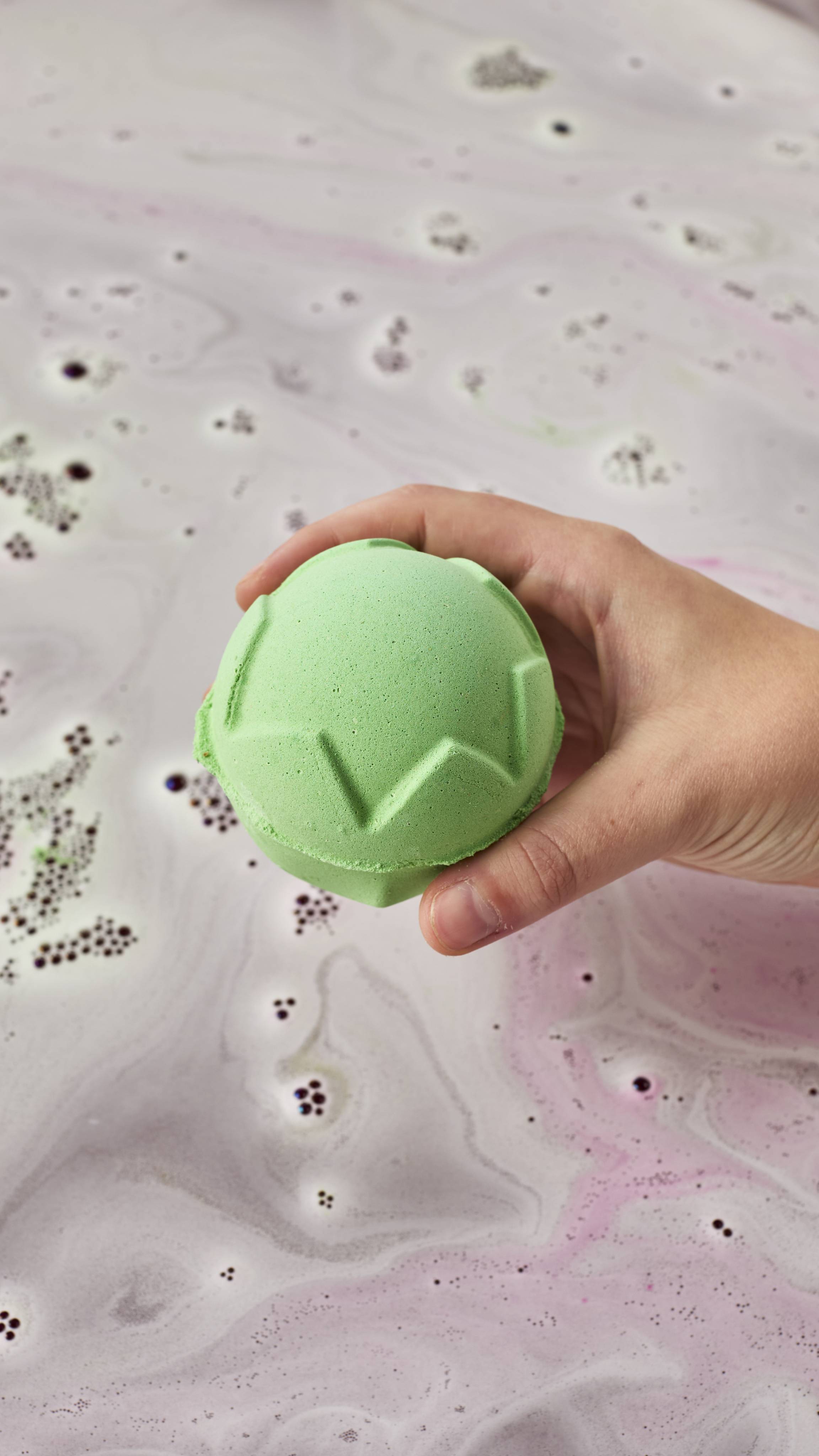 A hand holds a spherical light-green "Lord Of Misrule" bath bomb with carved geometric patterns over bathwater swirling with pale lavender foam and delicate bubbles.
