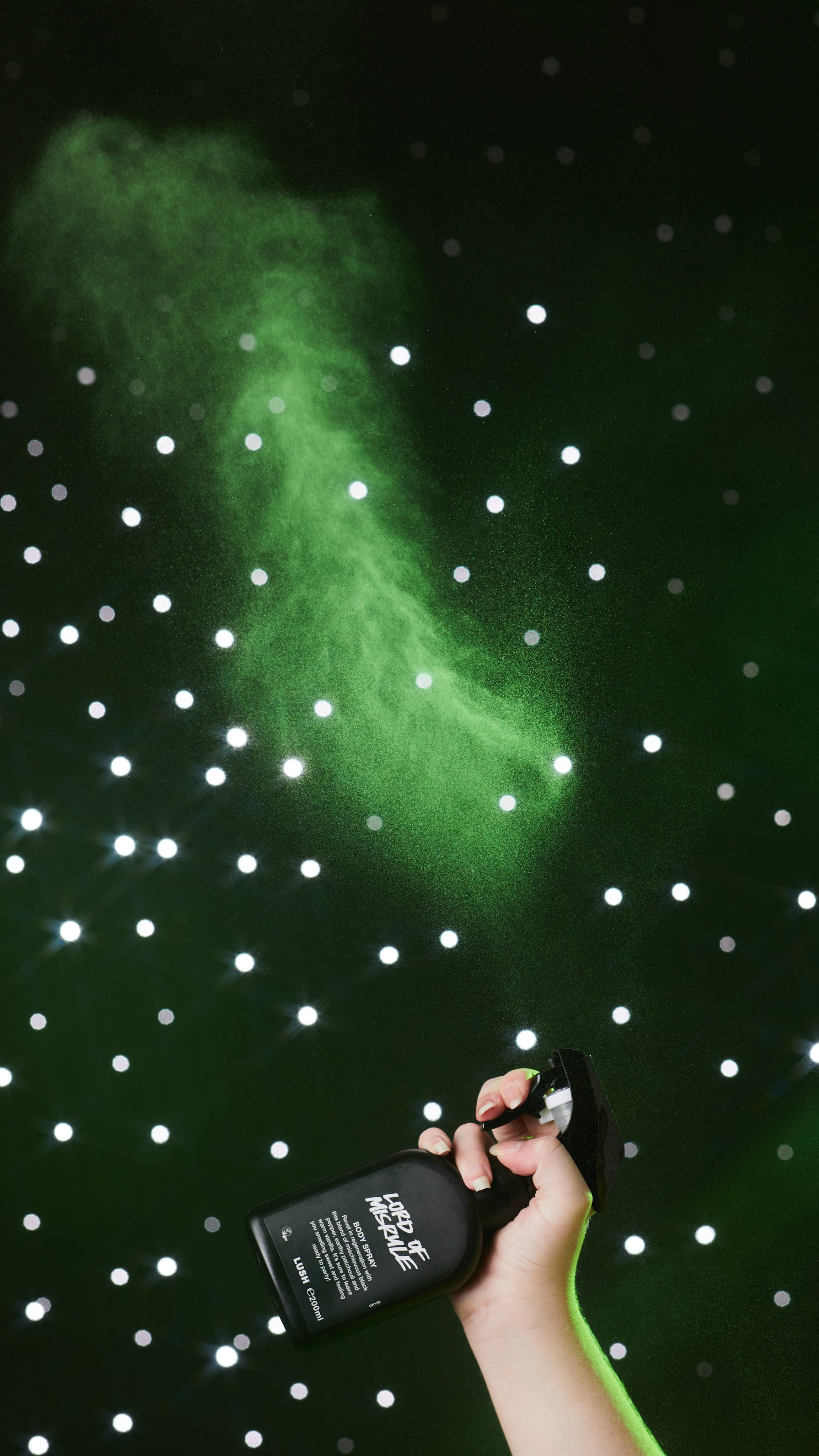 Image shows a close-up of models hand as they spray the bottle in the air, misting in front of a black, twinkling background. 