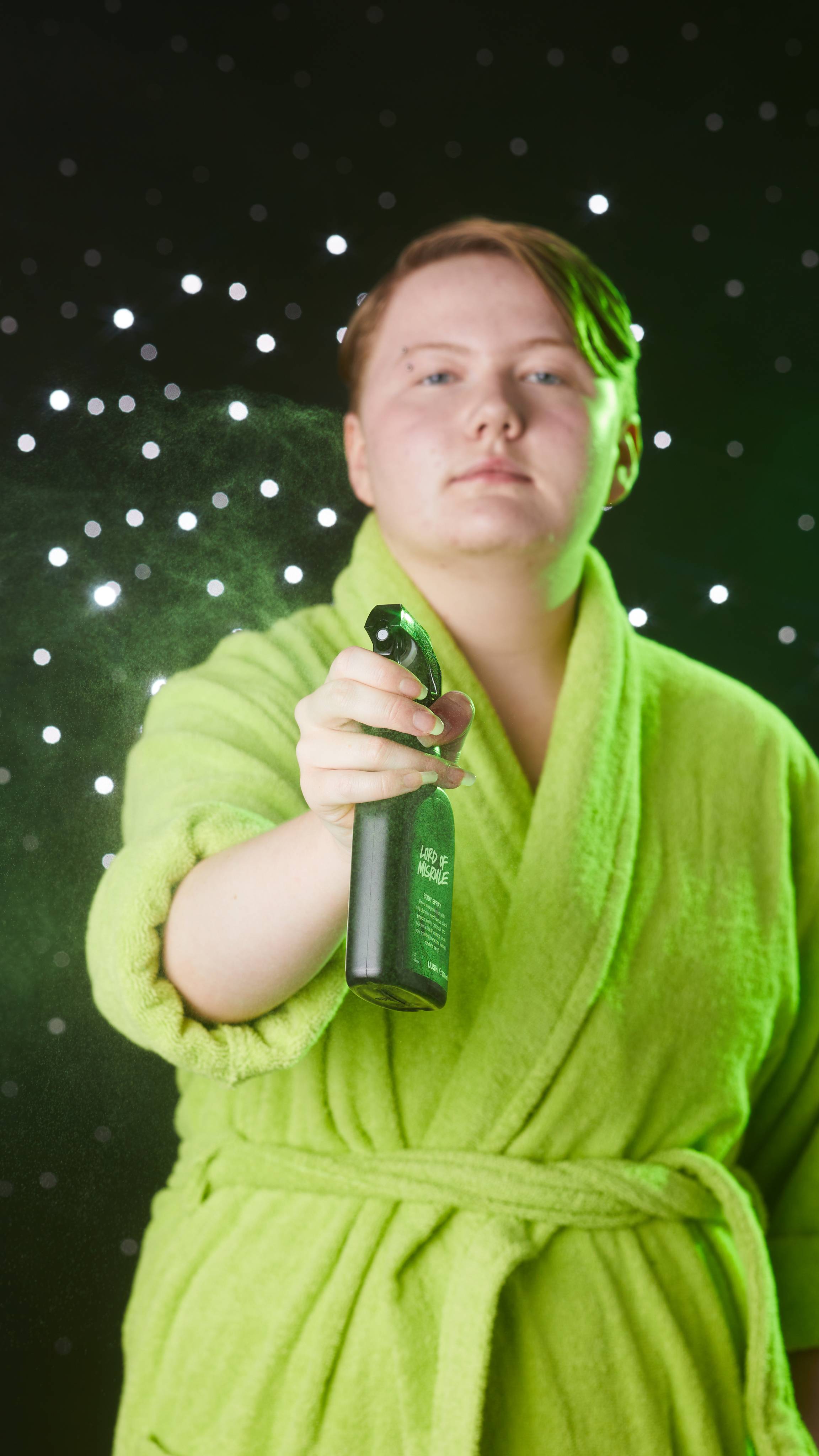 Model is stood on a black, twinkling background in a neon green robe spraying the bottle towards the camera.