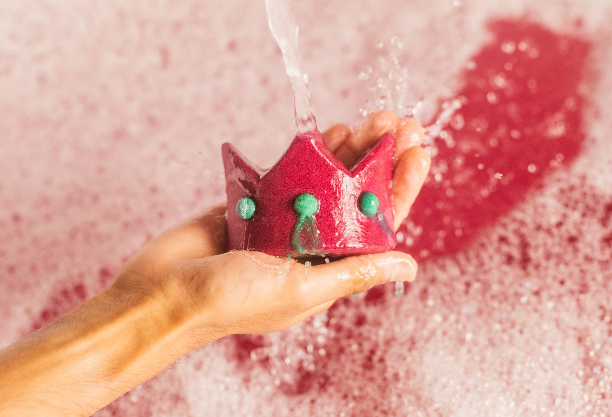 Image shows a close-up of bubble bar being held under running tap water. There is deep pink bathwater covered in bubbles behind.