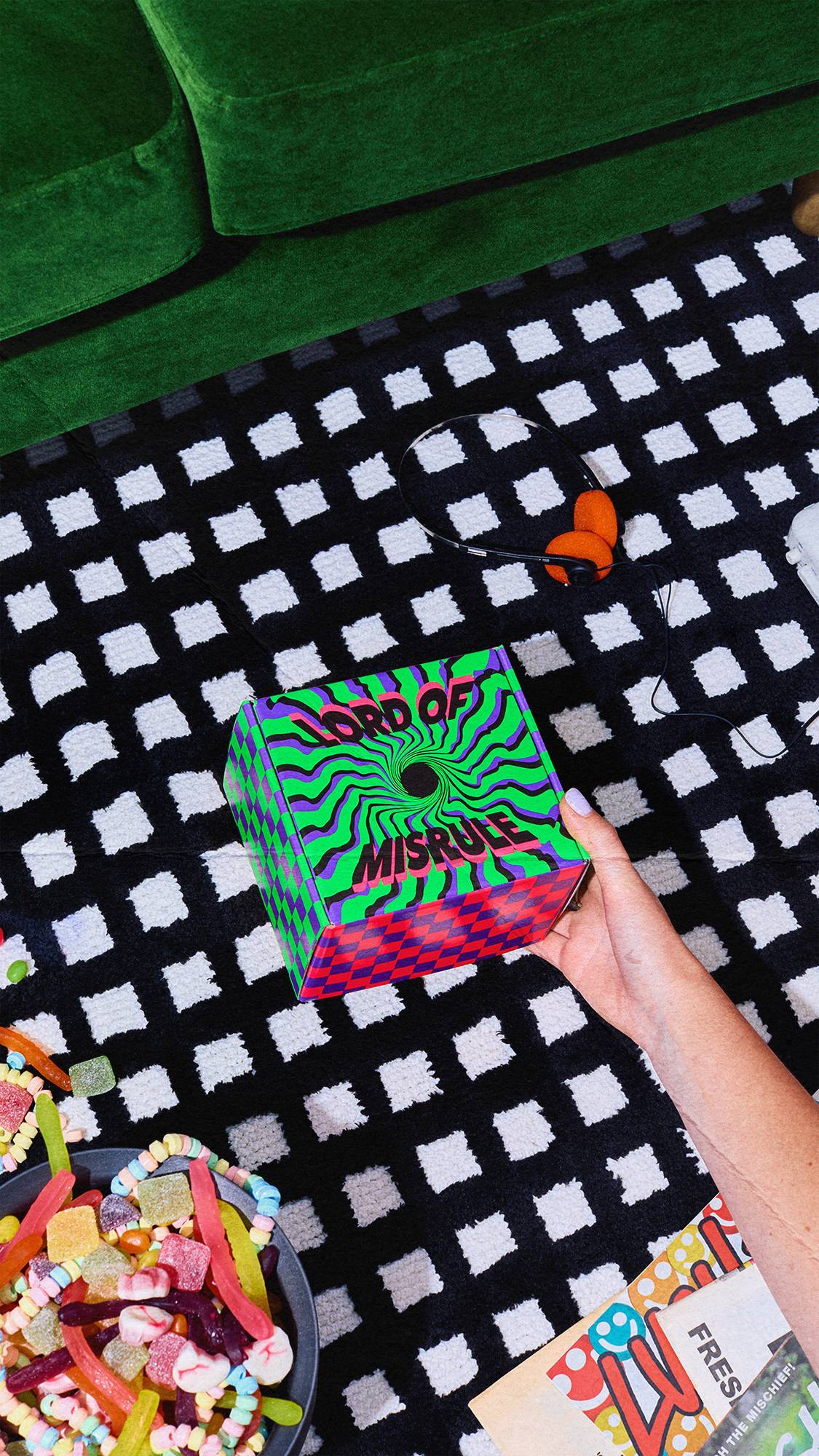 A hand reaches down to hold the psychedelic green and purple Lord of Misrule gift box on a monochrome checkered rug, next to a bowl of colourful sweets, retro headphones, and a green velvet sofa.