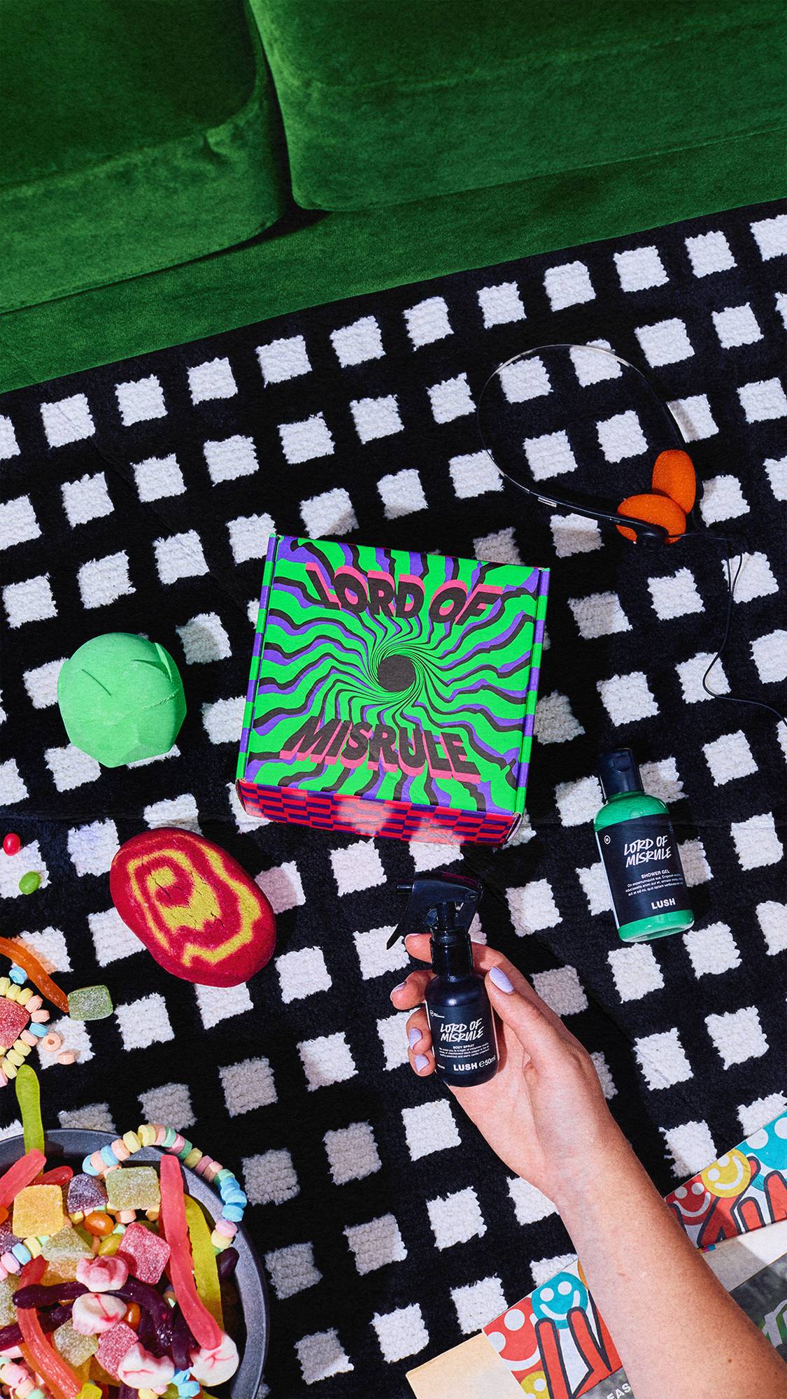 An aerial view of the Lush Lord of Misrule gift set arranged on a monochrome checkered rug. A hand holds the body spray alongside a green shower gel, bath bomb, swirled bubble bar, headphones, and a bowl of sweets.