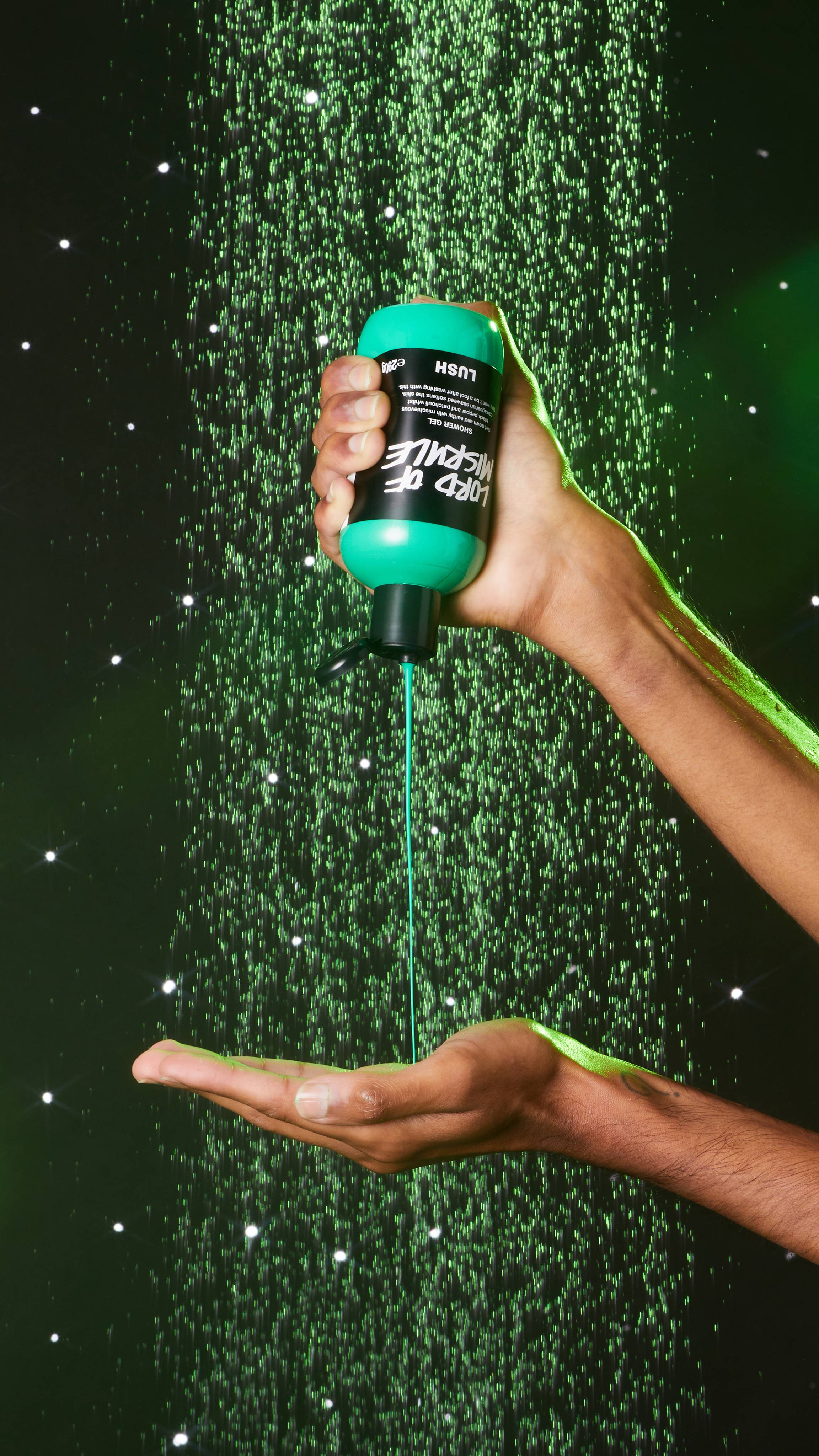  Image shows a close-up of the model squeezing the shower gel from the bottle into their hand on an emerald-green background.