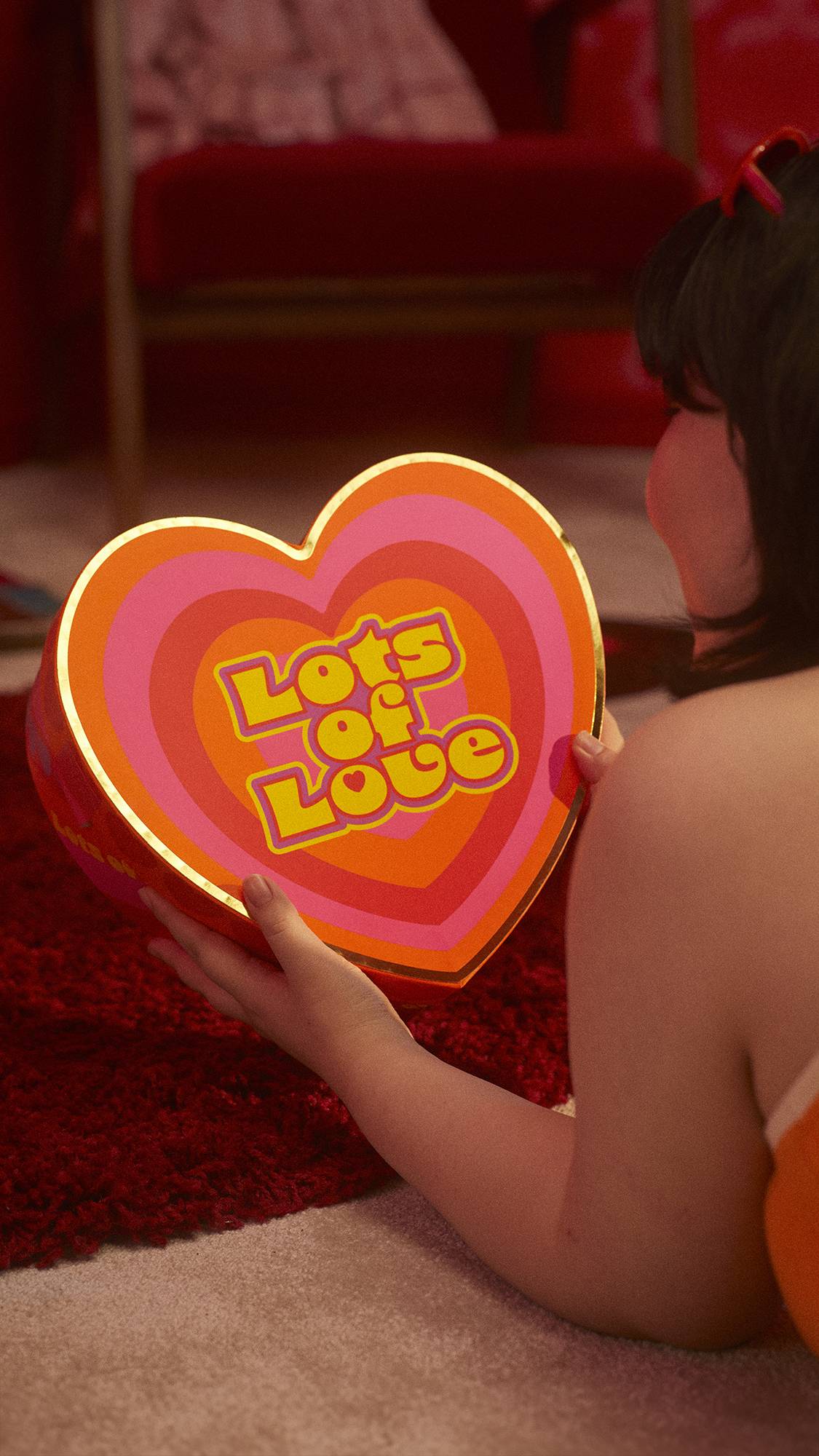 The image shows the model facing away as they are lying on the floor and holding the Lots of Love gift box. 