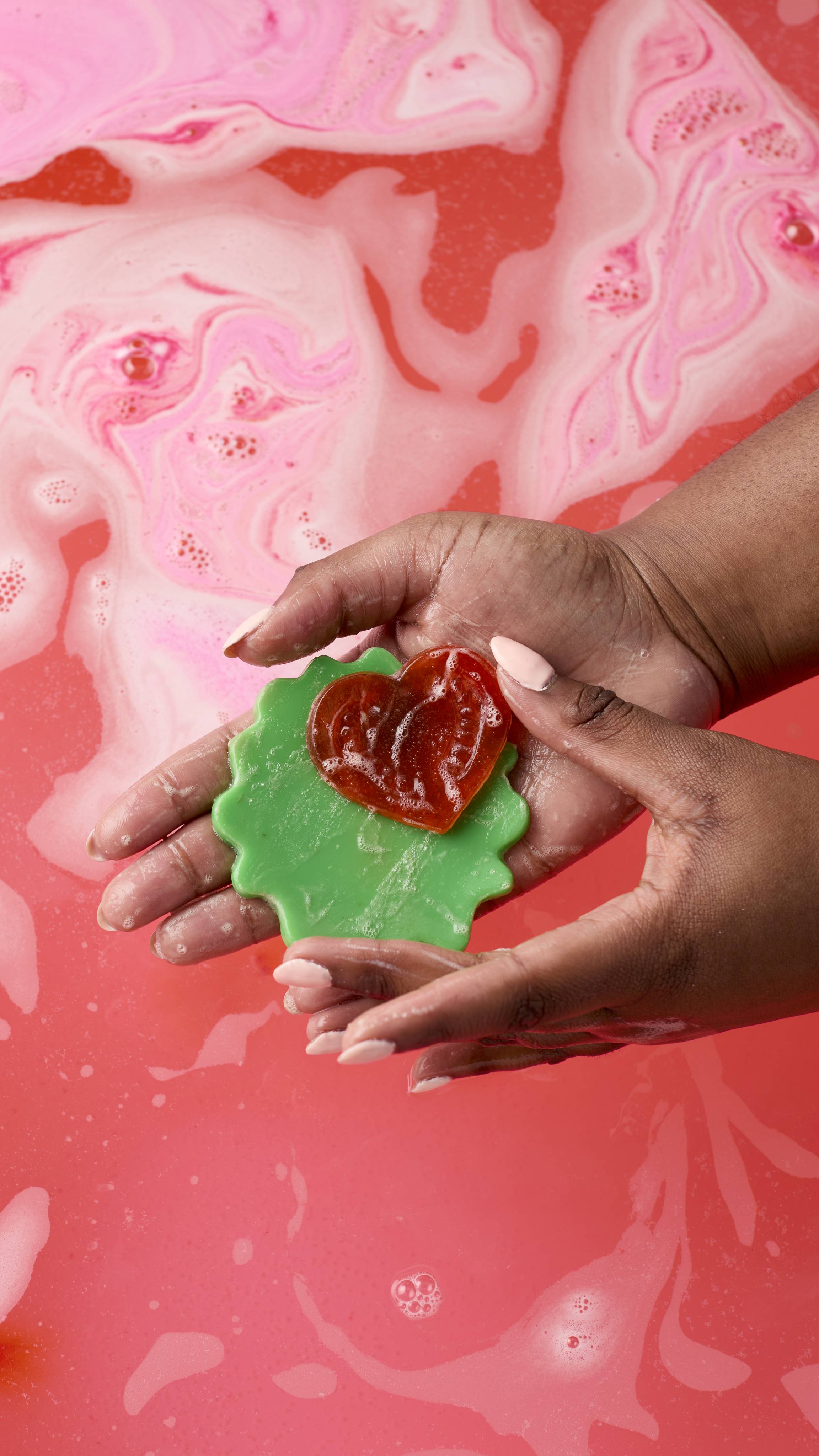 A person with dark skin holds a green, scalloped-edged lettuce soap and a small red heart-shaped tomato jelly over pink, swirling bathwater. Their hands are damp, and the background shows bubbly, pastel pink patterns in the water.