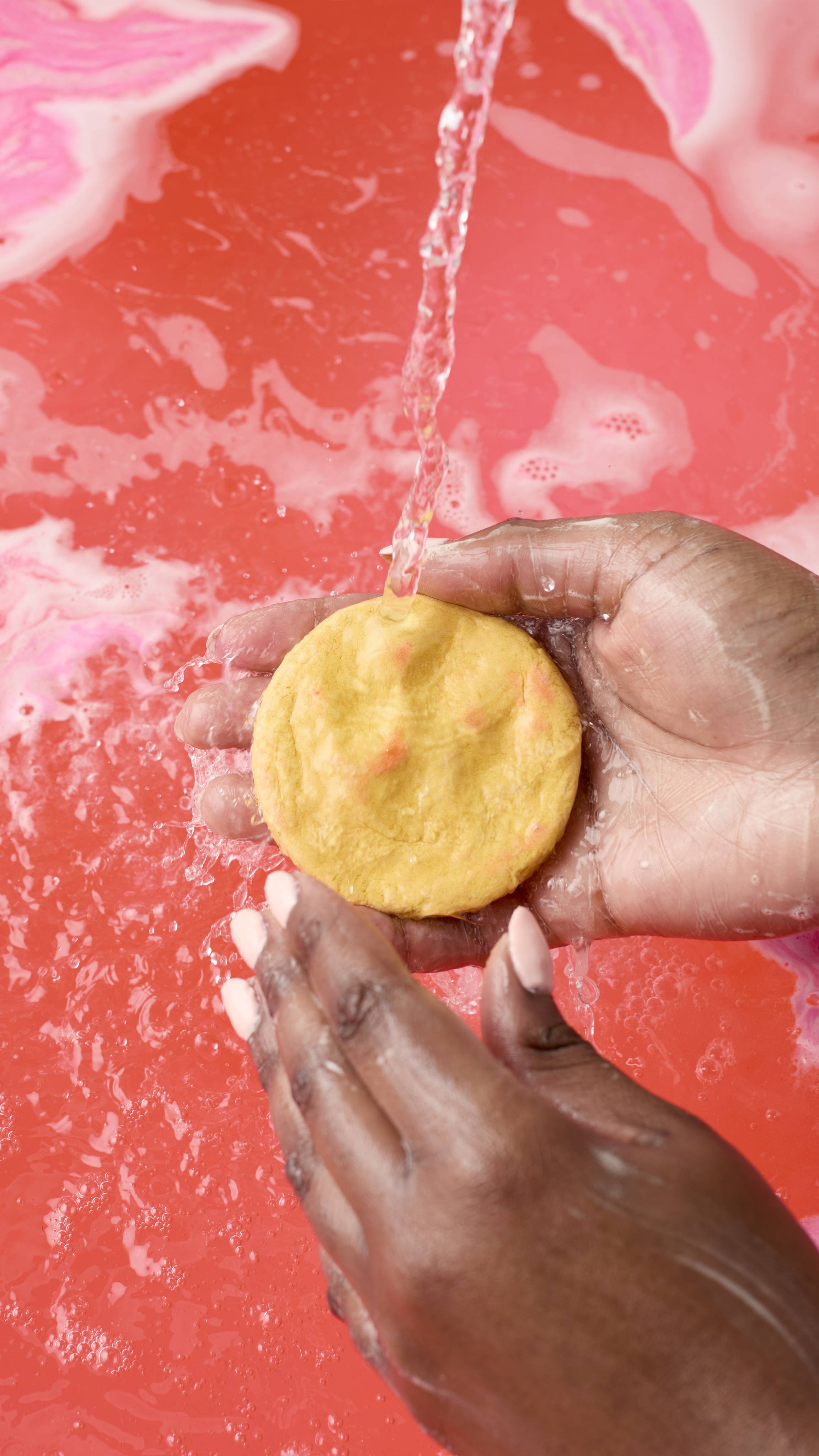 A person with dark skin holds a round, yellow burger bun-shaped bubble bar under a stream of running water. The water splashes against the bar and their hands, set against a backdrop of vibrant, swirling pink bathwater
