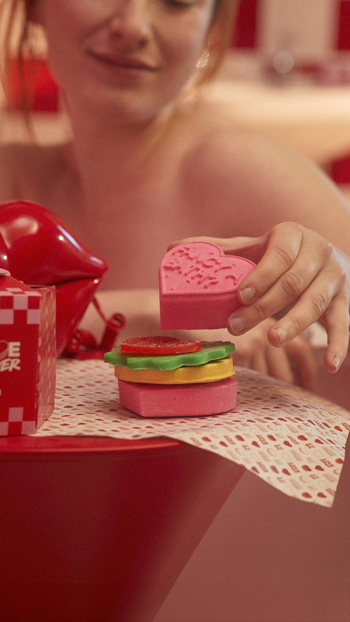 Image shows the Love Burger sat on a table in a red-themed bathroom as the model lifts off the top bun/bath bomb product.
