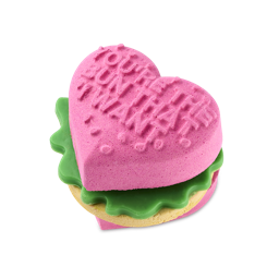 Love Burger. A playful bath bomb shaped like a burger with heart-shaped buns. The top bun is bright pink with embossed text reading "You're The Bun That I Want", layered over green soap "lettuce" and a beige "patty" bubble bar.