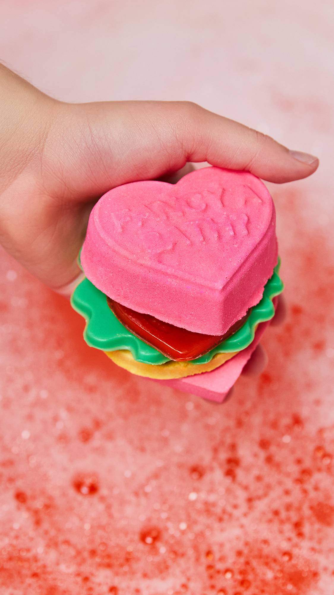 Model is holding all Love Burger products together, formed of two burger bun bath bombs, tomato and lettuce soap, and patty bubble bar.