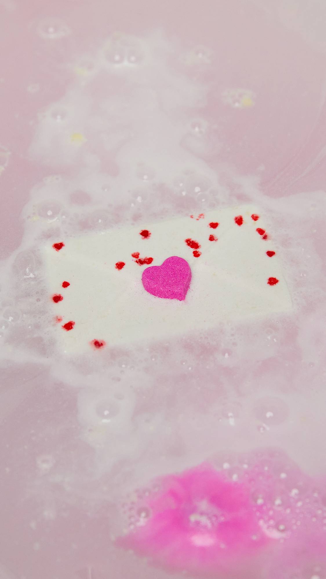 The Love Letter bath bomb dissolves in the bath water, slowly releasing delicate swirls of pale pink and white 
