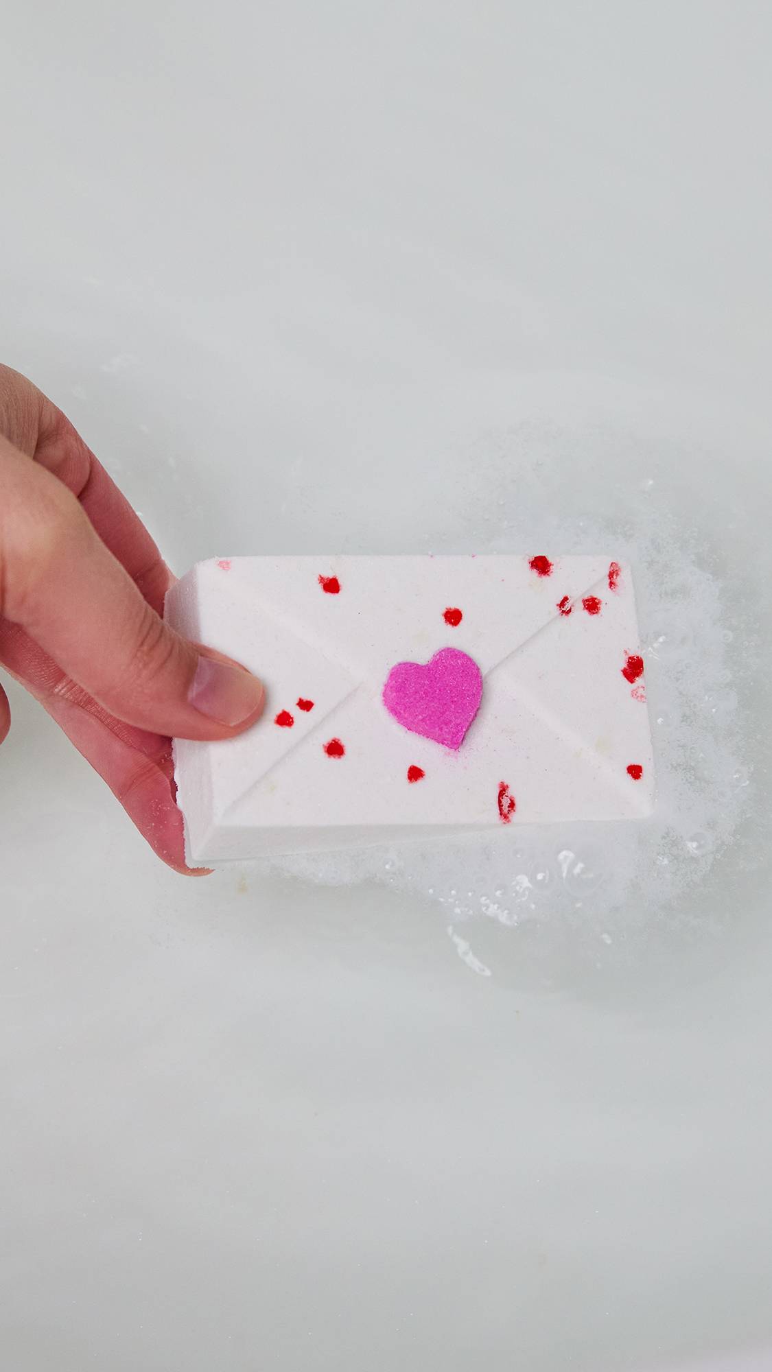 The image is a close-up of the model's fingertips holding the Love Letter bath bomb as they are about to drop it into the water. 