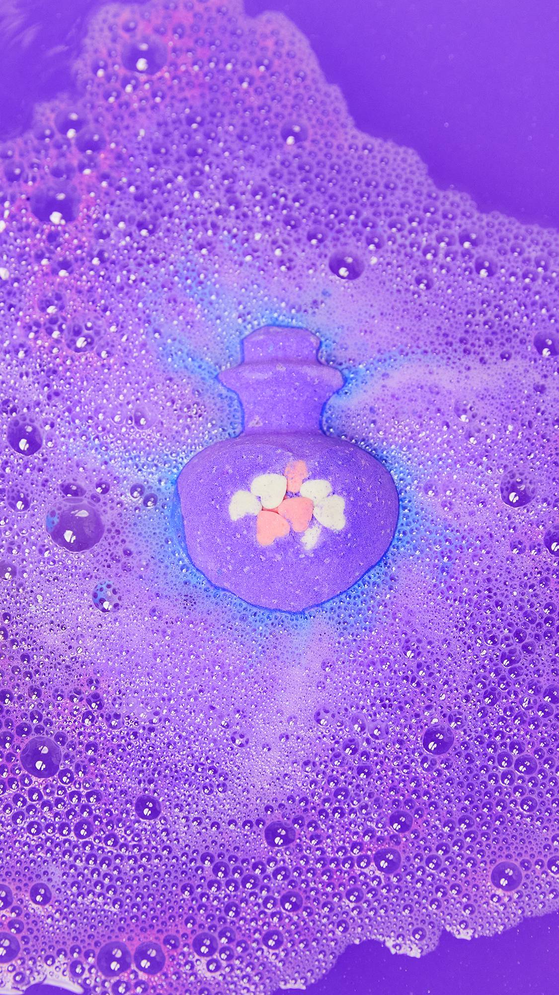 Love Potion bath bomb sits in the middle of the water, the pale pink and white heart confetti just visible, surrounded by deep purple.