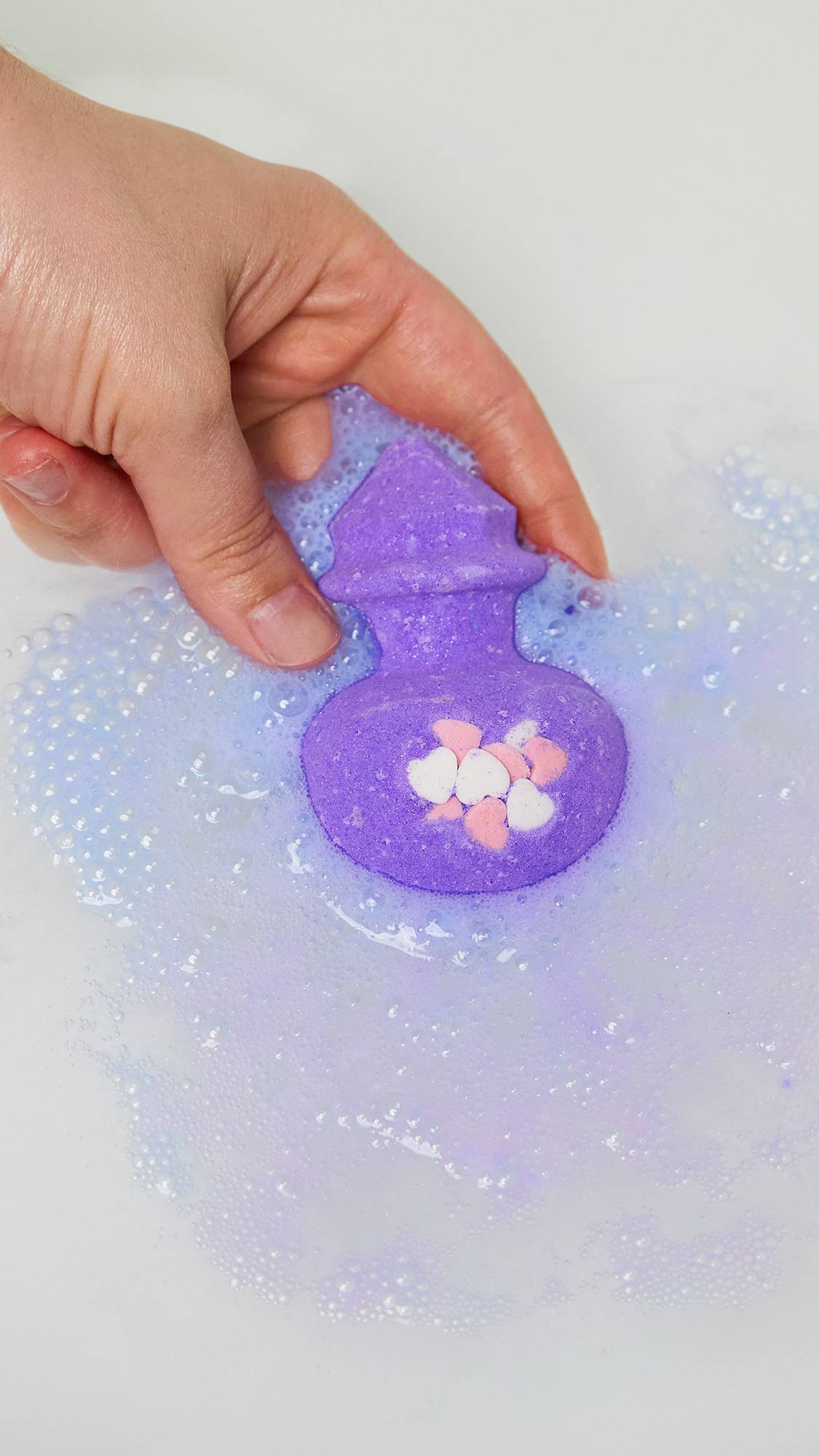 The image shows a close-up of the model's hand as they are gently placing the Love Potion bath bomb into the water. 
