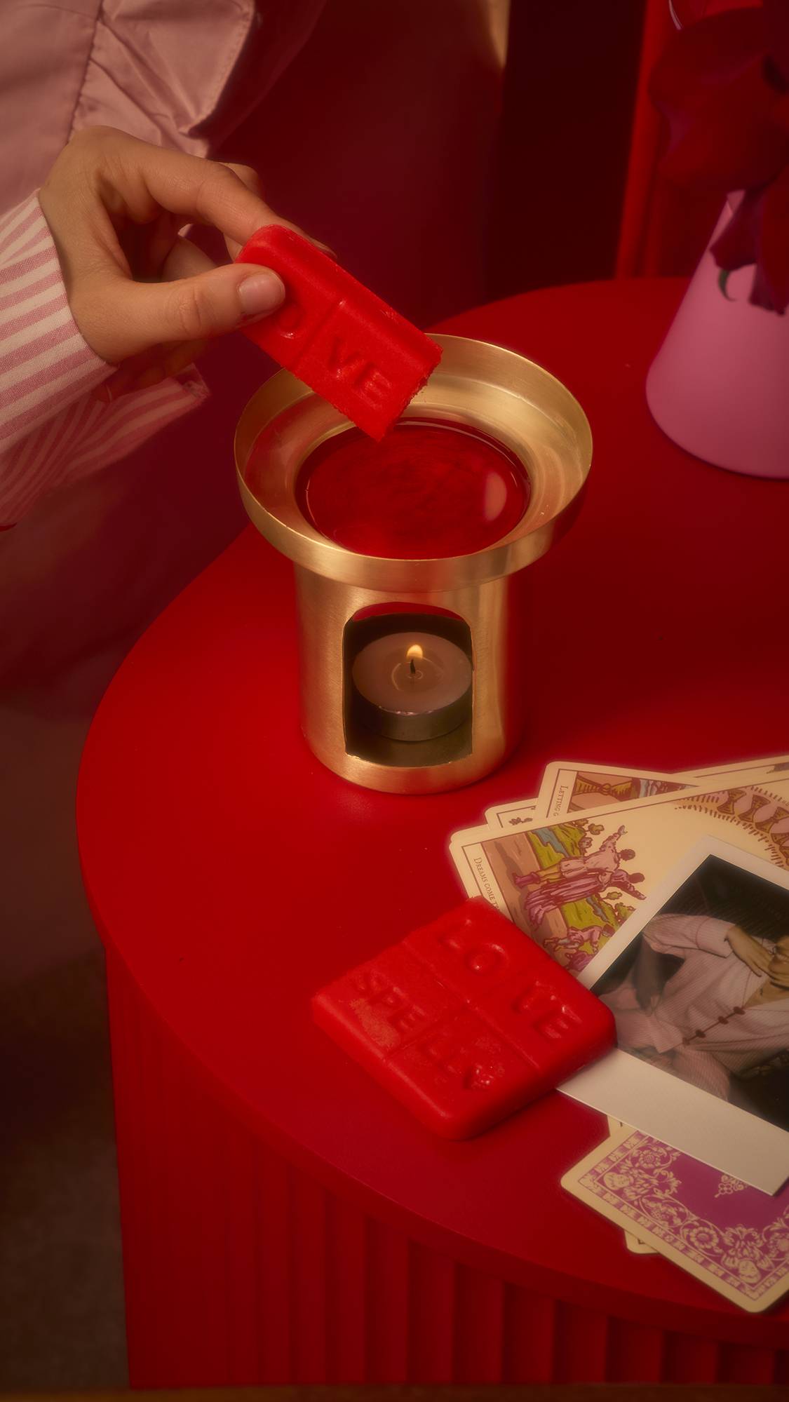 A rectangular red bar embossed with "LOVE SPELL" and heart icons, designed to be broken into squares for use in a wax warmer or bath. The image shows the model placing two squares of the Lush Melt into a golden wax warmer. 