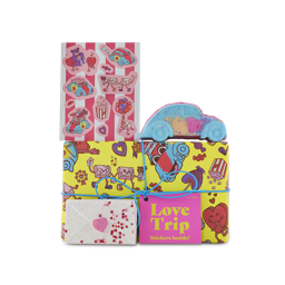 Love Trip. A gift box wrapped in yellow paper decorated with Valentine-themed illustrations and tied with a blue string. The car-shaped Love Bug bath bomb and Love Letter bath bomb sit around the box with a sheet of Valentine's Day stickers. 