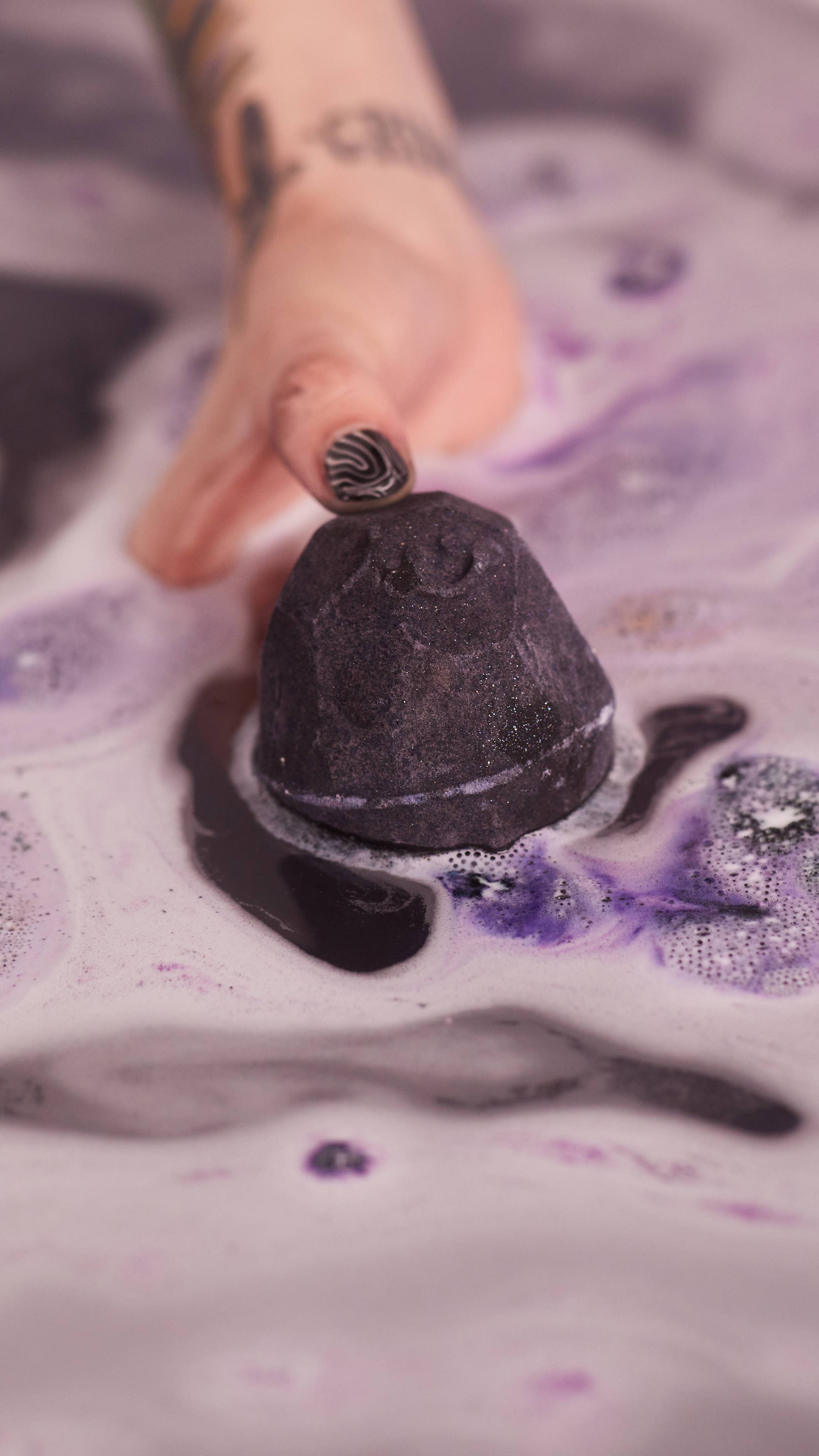 A close-up shot of model holding the bath bomb half out of water. which has a deep, velvety, dark purple colour.