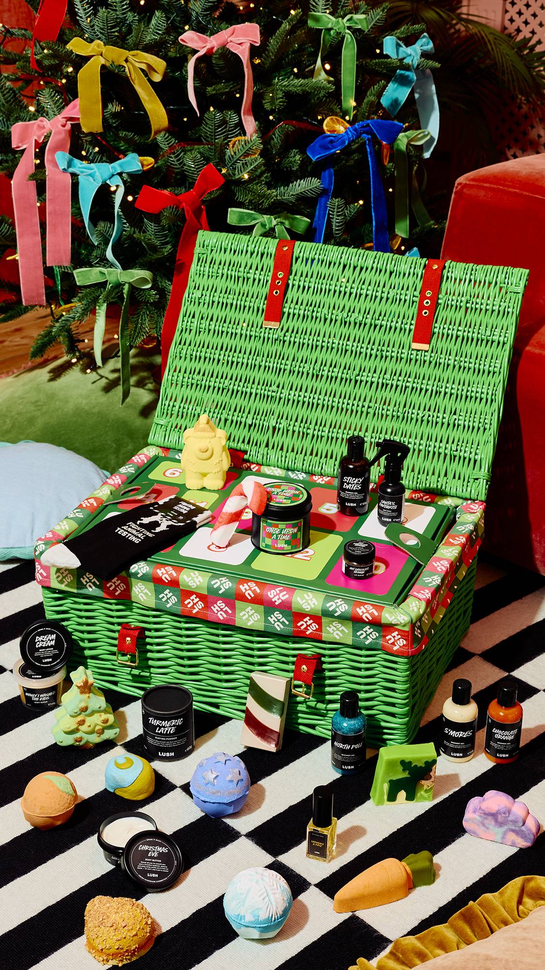 An open green hamper with red straps displays festive Lush treats, surrounded by Christmas bath bombs, soaps, and bottles scattered on a striped rug before a Christmas tree adorned with colourful bows.