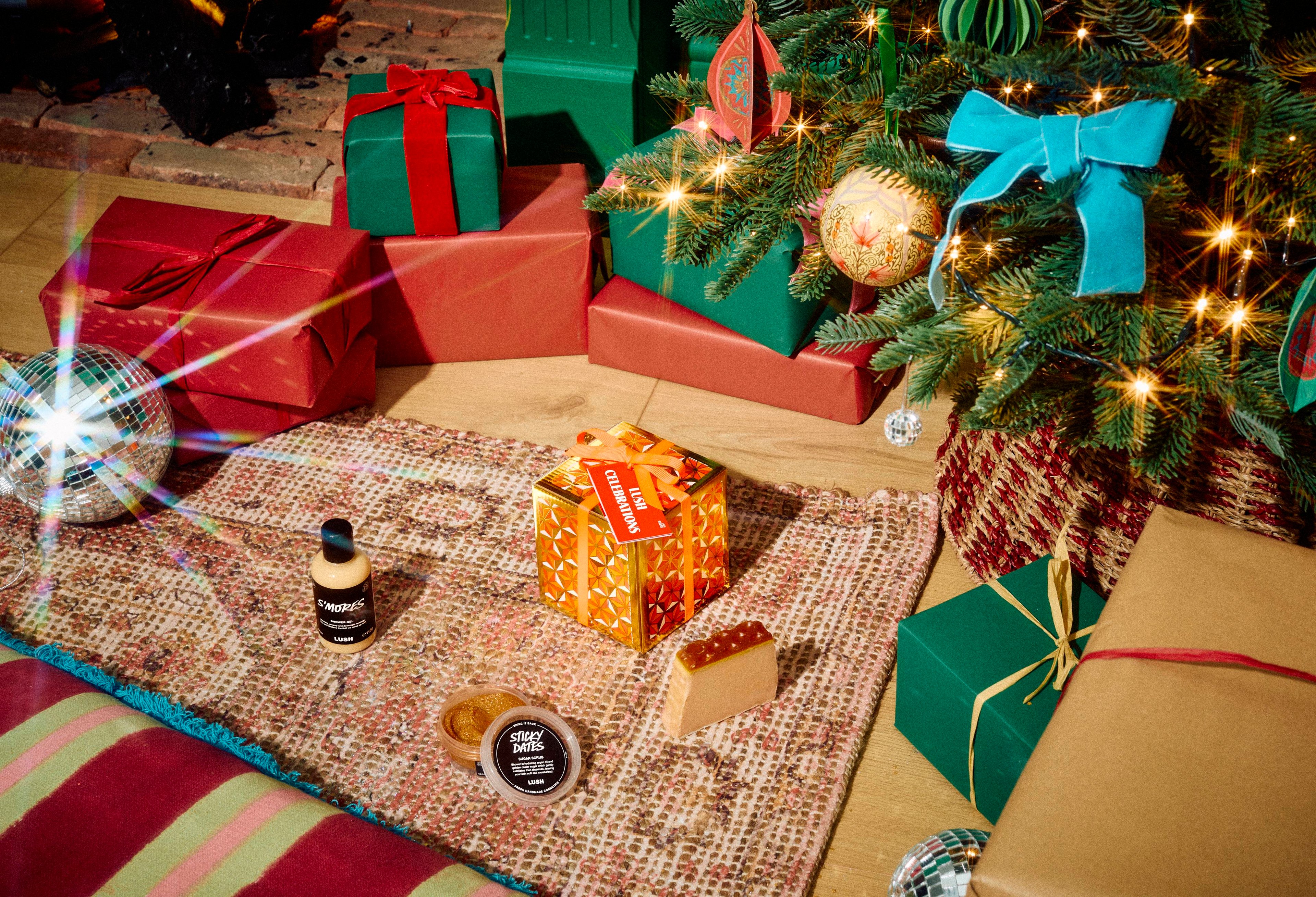 The Lush Celebrations gift box sits on a patterned rug with S'mores shower gel, Sticky Dates body scrub, and a slice of soap, surrounded by wrapped presents under a decorated Christmas tree.