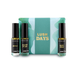 The image shows a "LUSH DAYS" perfume gift set. Three cylindrical glass perfume bottles are arranged around a vibrant teal box with a wavy pattern and yellow text. Perfumes include 'Grass', 'Breath of God' and 'Dear John'.