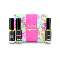 The image shows a "LUSH Romance" perfume gift set. Three thin glass perfume bottles with amber and clear liquids are arranged around a light blue box with vibrant floral patterns and a bright pink band.