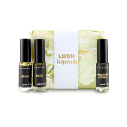 The image shows a "LUSH Triptych" perfume gift set. Three thin, cylindrical glass perfume bottles with amber and clear liquids are arranged around a pale yellow and green marbled box with a white band.