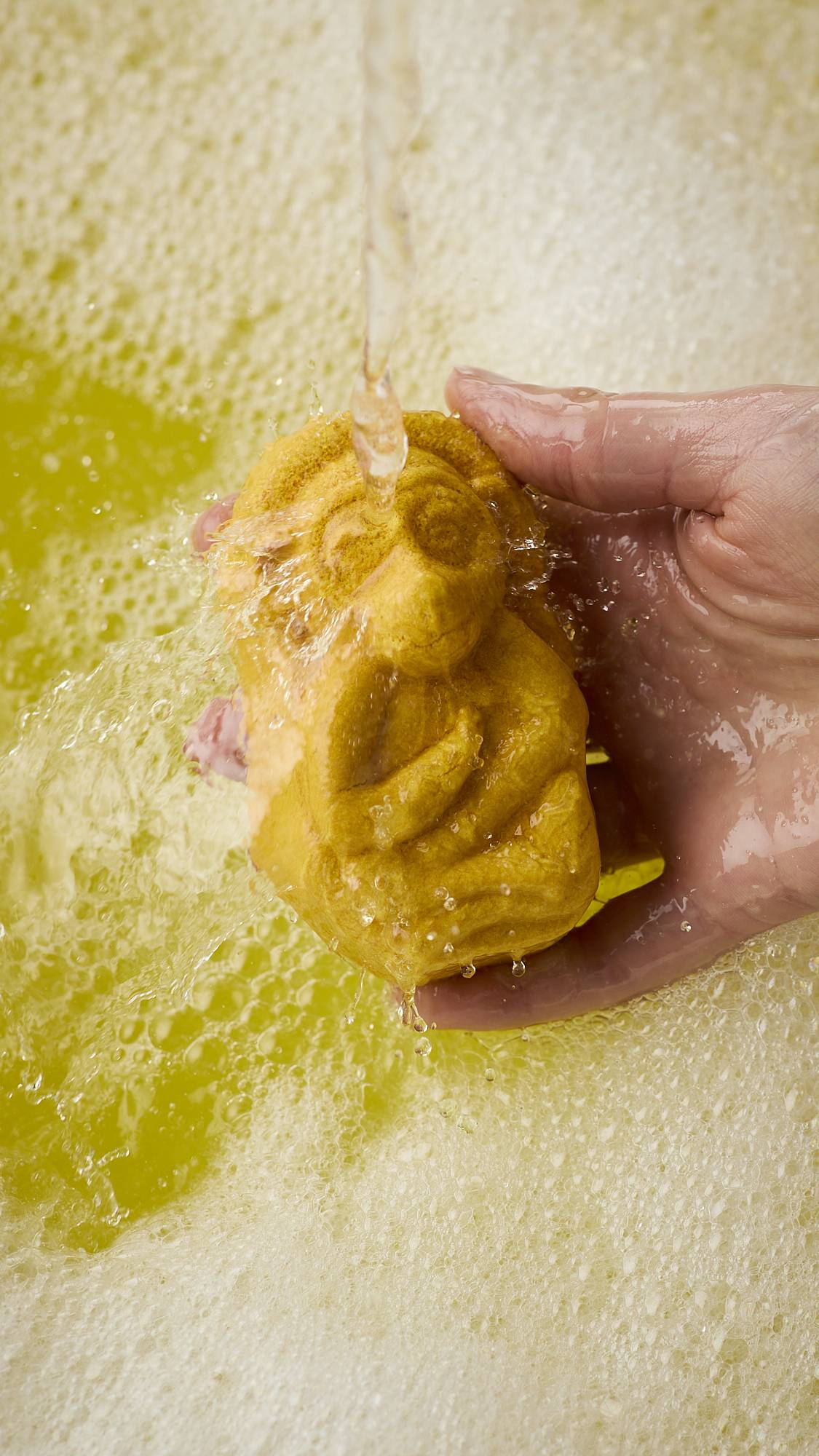 A close-up image of the model's right hand holding the caramel-coloured, Macaque monkey-shaped bubble bar under running bath water. The product creates bright, sunshine-yellow water and a thick blanket of bubbles. 