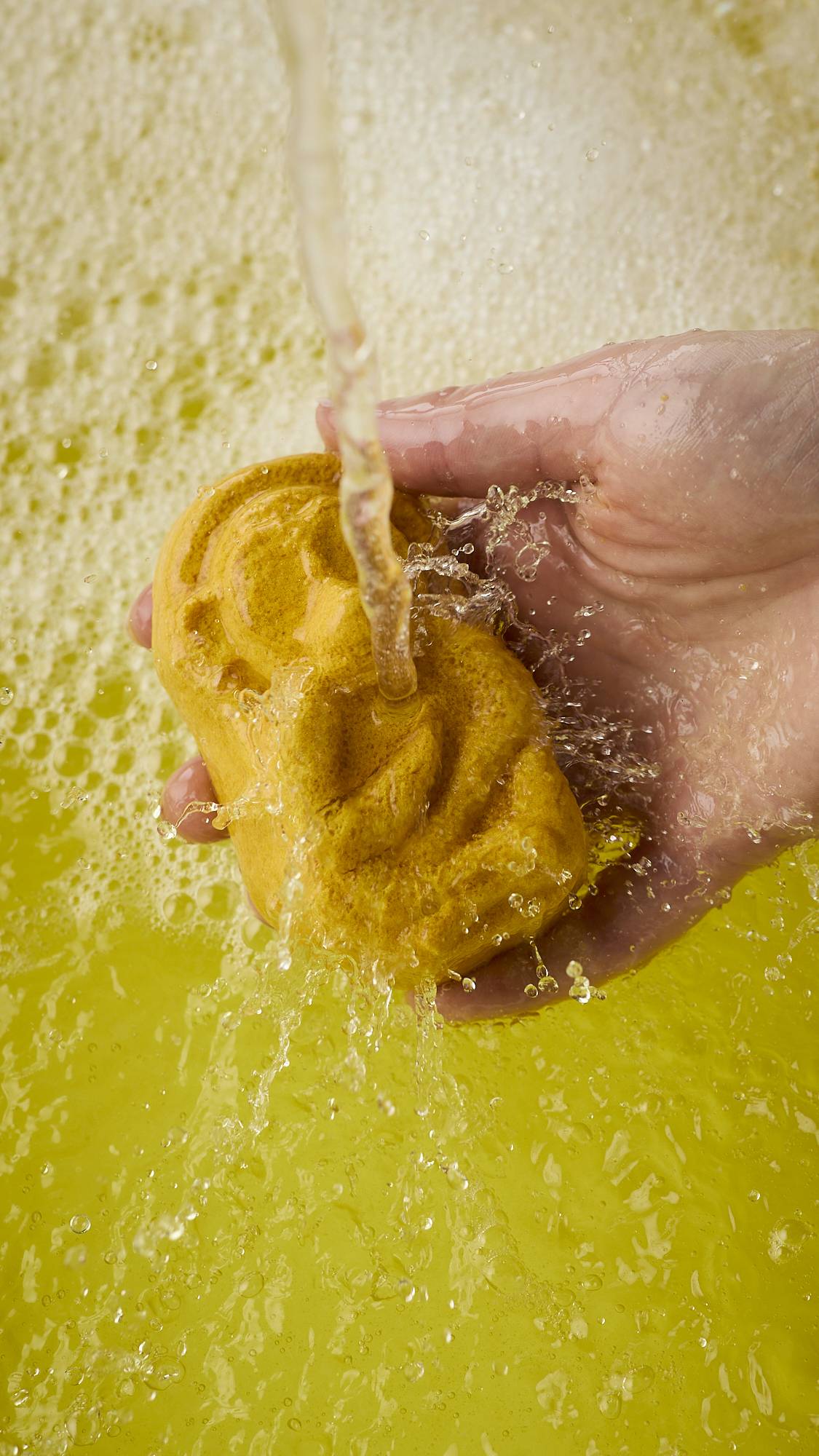 A close-up image of the model's right hand holding the caramel-coloured, Macaque monkey-shaped bubble bar under running bath water. The product creates bright, sunshine-yellow water and a thick blanket of bubbles. 