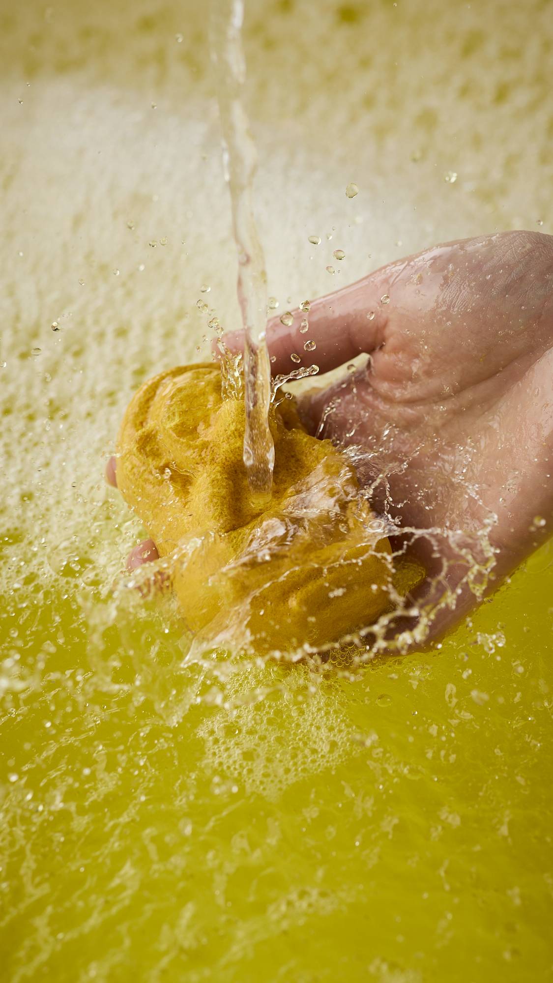 A close-up image of the model's right hand holding the caramel-coloured, Macaque monkey-shaped bubble bar under running bath water. The product creates bright, sunshine-yellow water and a thick blanket of bubbles. 