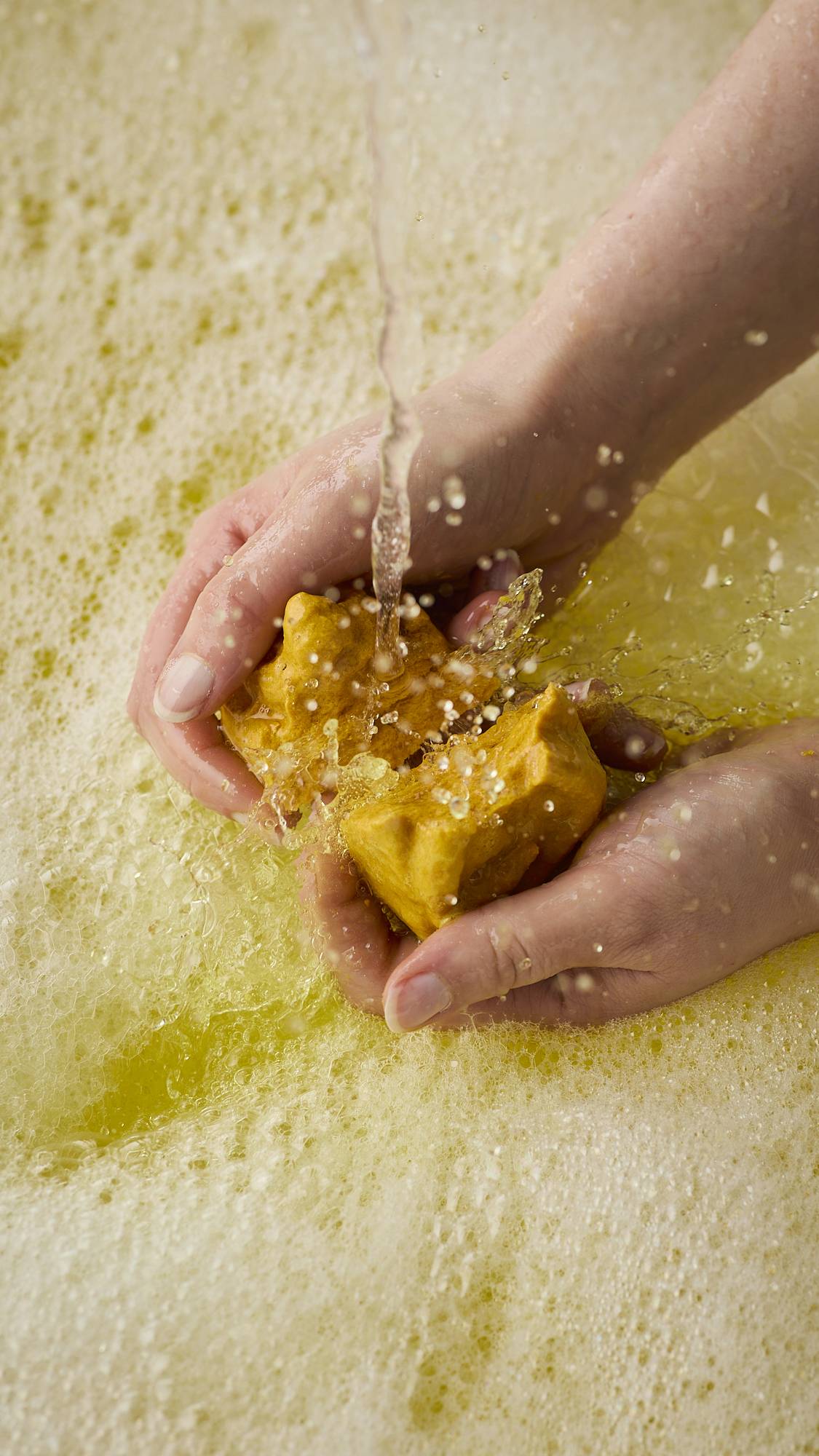 A close-up image of the model's hands breaking and crumbling the caramel-coloured, Macaque monkey-shaped bubble bar under running bath water. The product creates bright, sunshine-yellow water and a thick blanket of bubbles. 