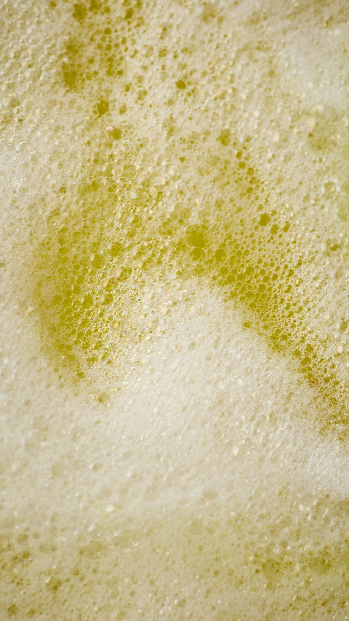 The image shows an aerial view of the bath after the model has crumbled the Macaque monkey-shaped bubble bar into their bath. The water is a vibrant sunshine-yellow and there is a thick blanket of white, foamy bubbles on top.