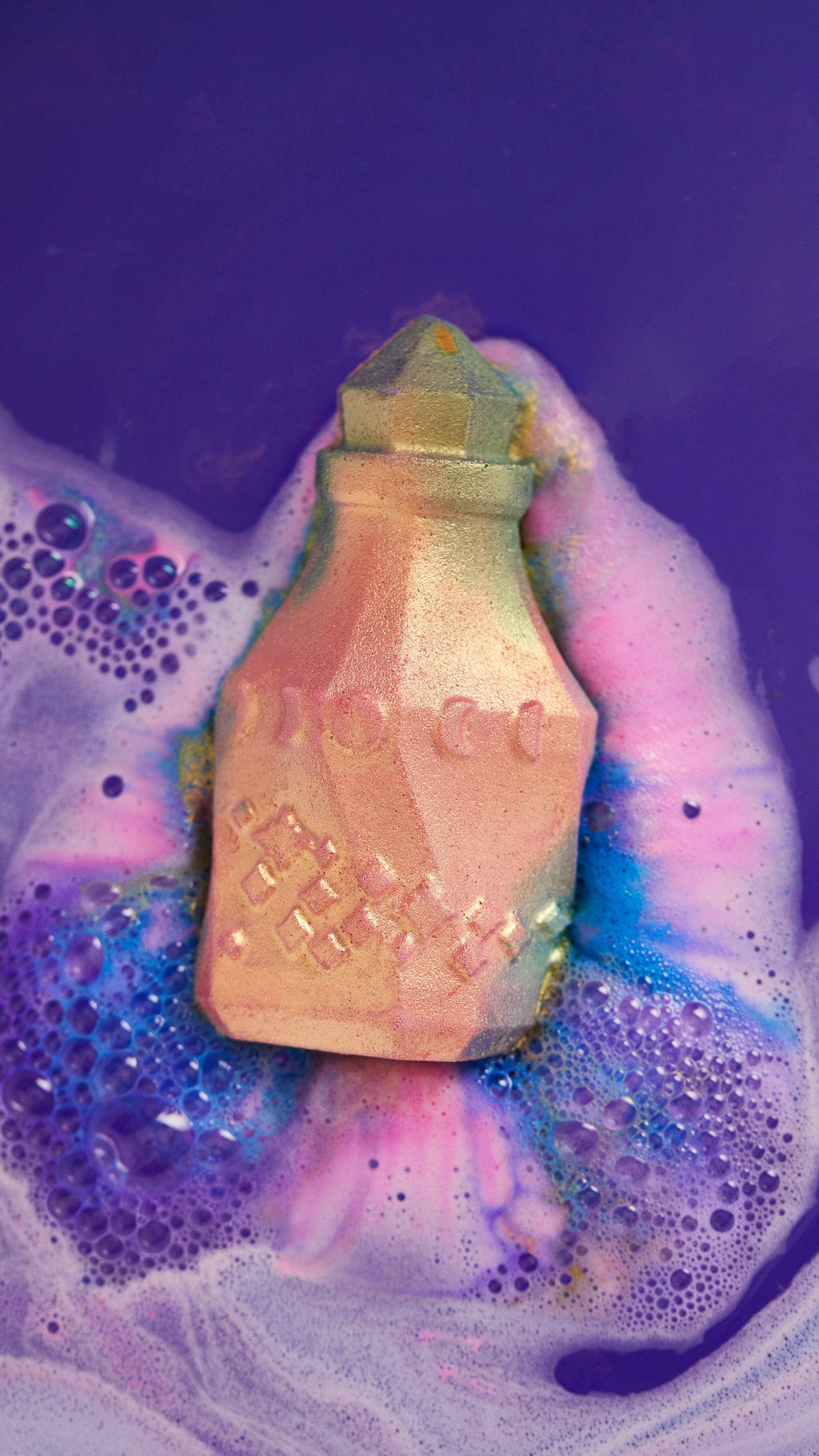 The image shows the Magic Potion bath bomb laying flat on top of the bath water as it begins to give off thick swirling ribbons of deep purples, pinks and blues. 