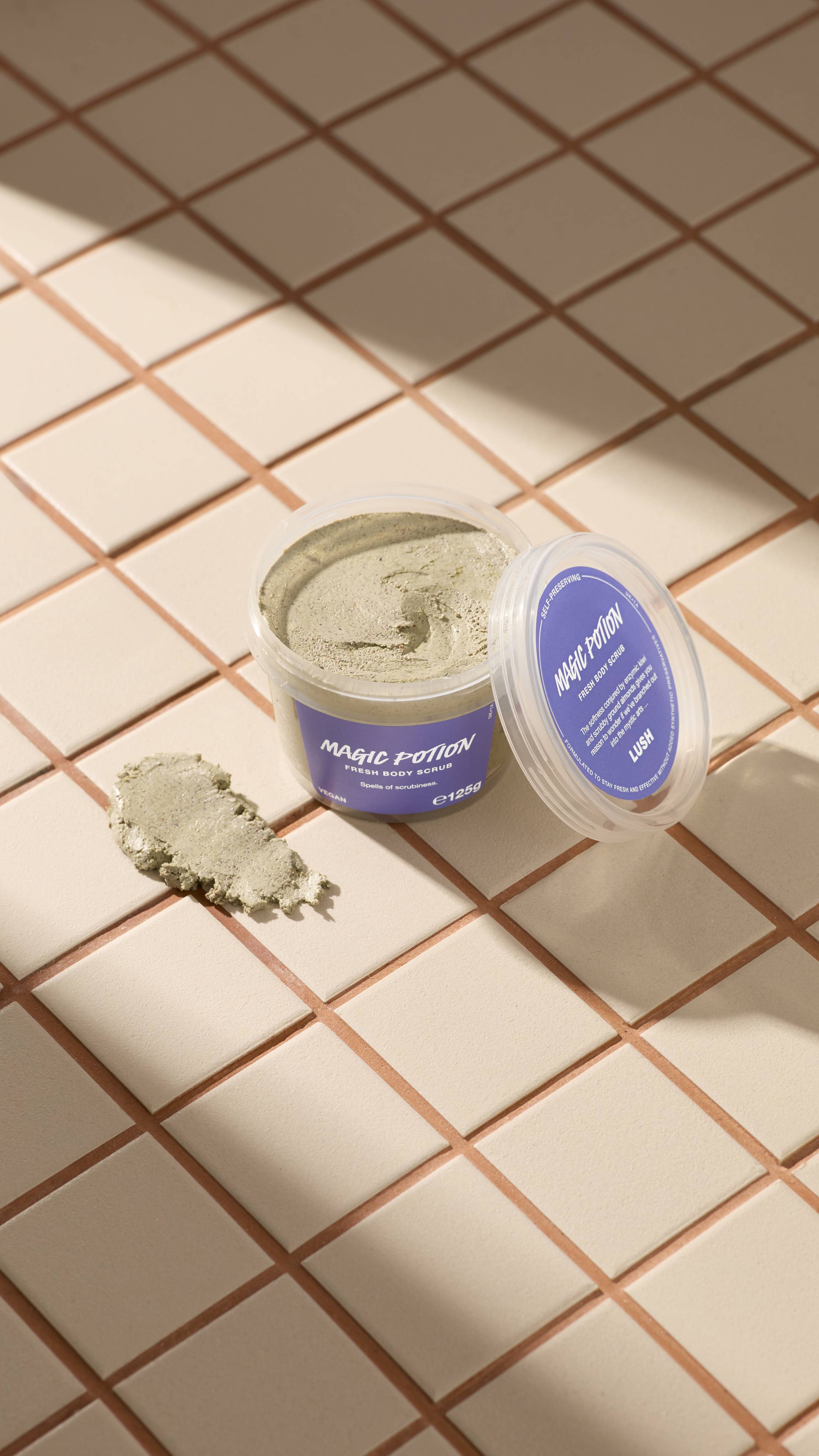 An open clear pot of Lush "Magic Potion Fresh Body Scrub" filled with pale sage-green paste sits on beige tiled surfaces beside its lid with a purple label, accompanied by a textured scoop of product.